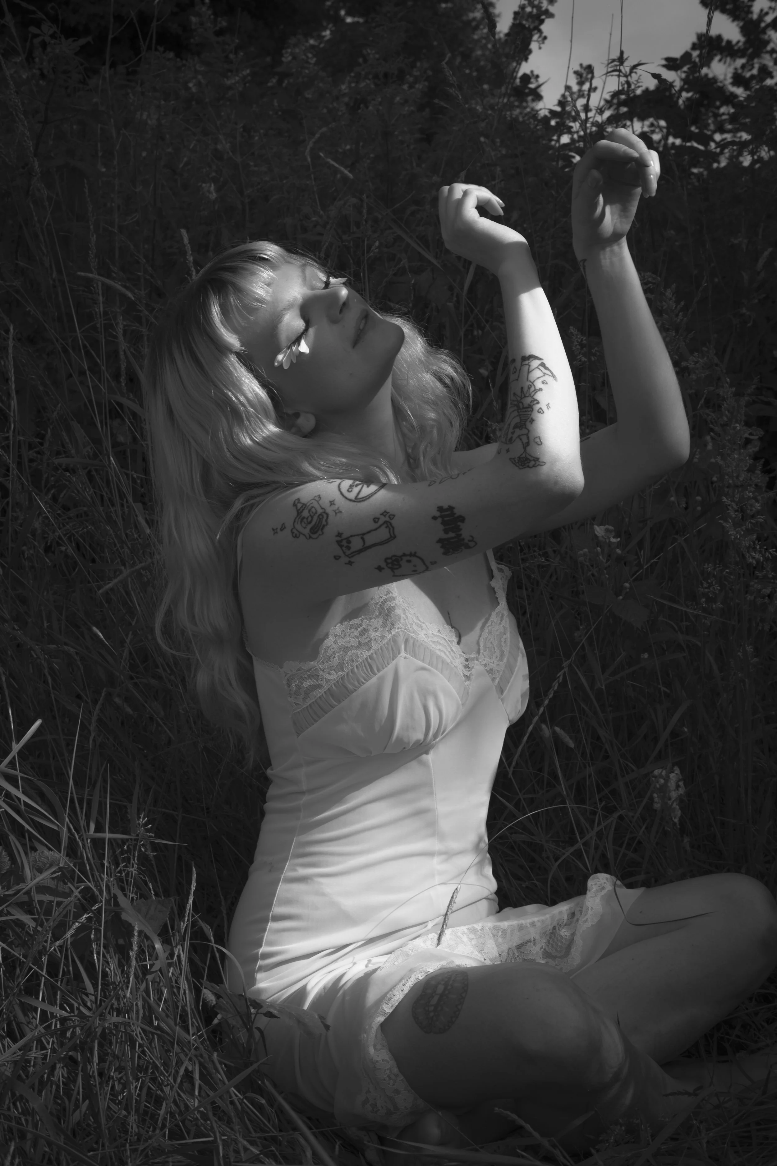 A woman with blonde hair and tattoos on her arms sitting in tall grass with closed eyes, smiling softly, in a natural outdoor setting. The photo is in black and white.