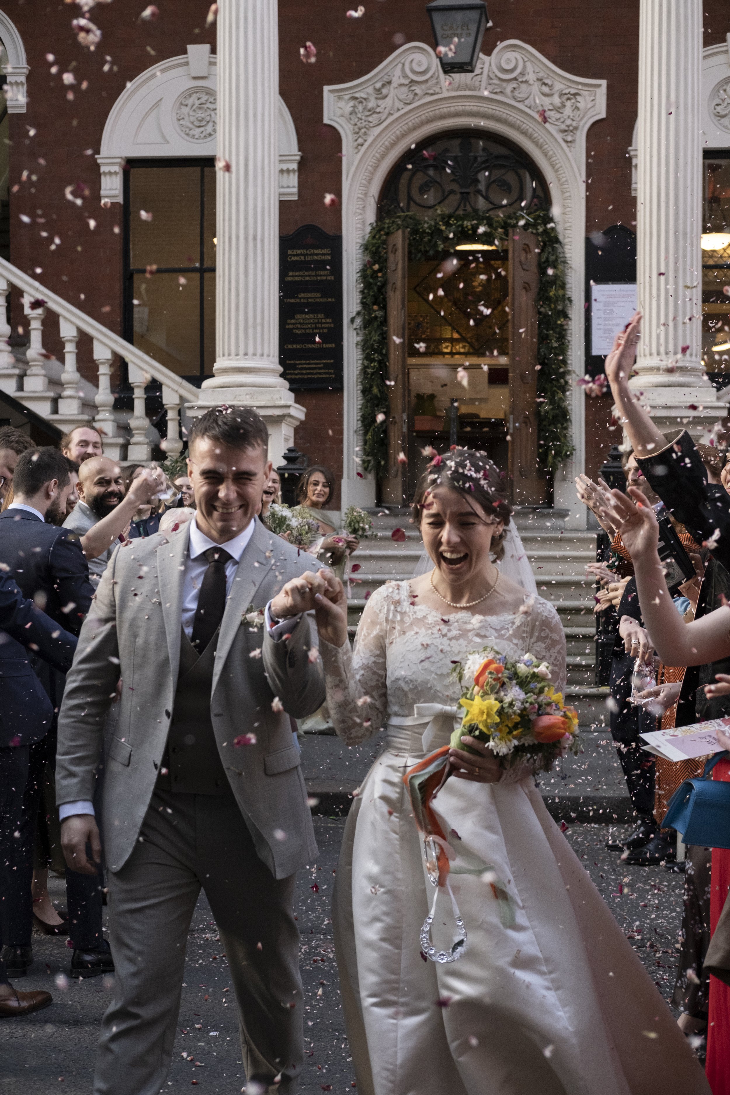 A bride and groom smiling as they walk hand in hand through a crowd of wedding guests throwing flower petals at a wedding celebration outside a church.