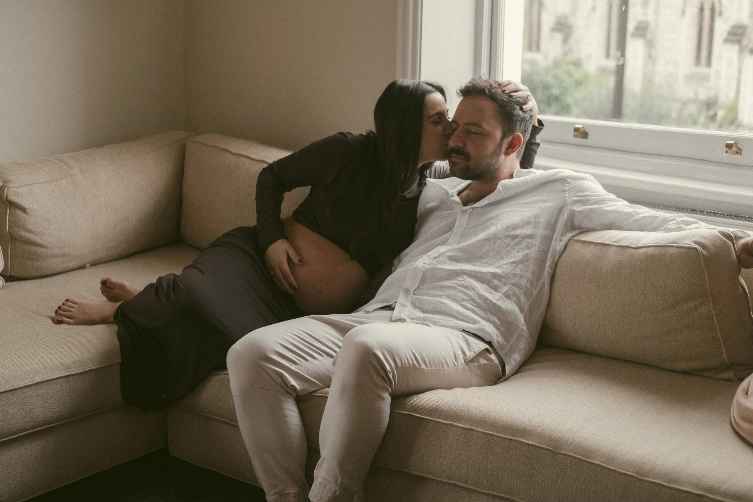 A woman leaning over to kiss a man on the forehead as he sits on a beige couch near a window.