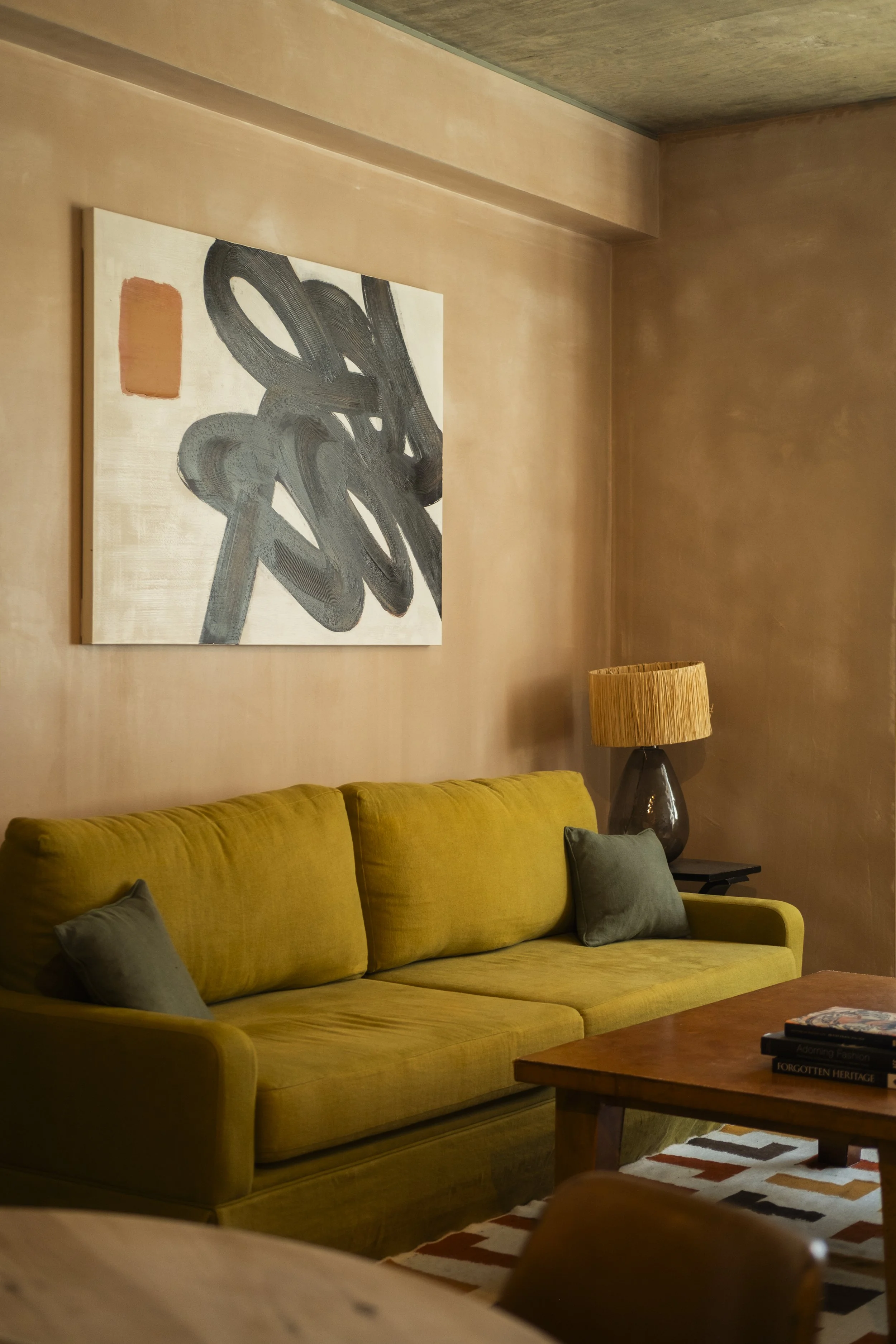 A yellow sofa with grey pillows, a wooden coffee table with books, a table lamp with a woven shade, a colorful area rug, and abstract art on a beige wall in a cozy living room.