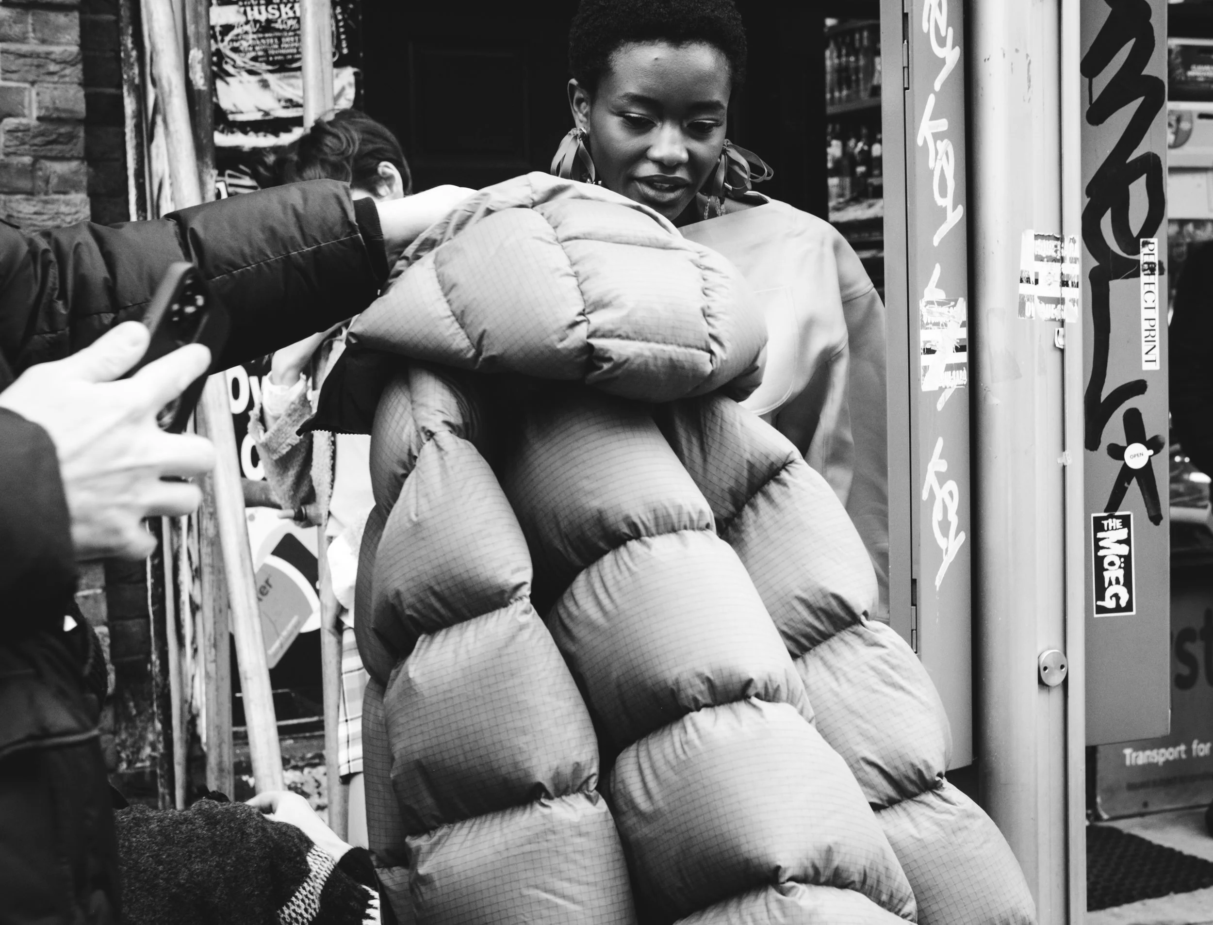 A woman with short curly hair and earrings stands on a city street, leaning on a railing, in front of a row of vending machines or kiosks, with a person taking her photo with a phone.