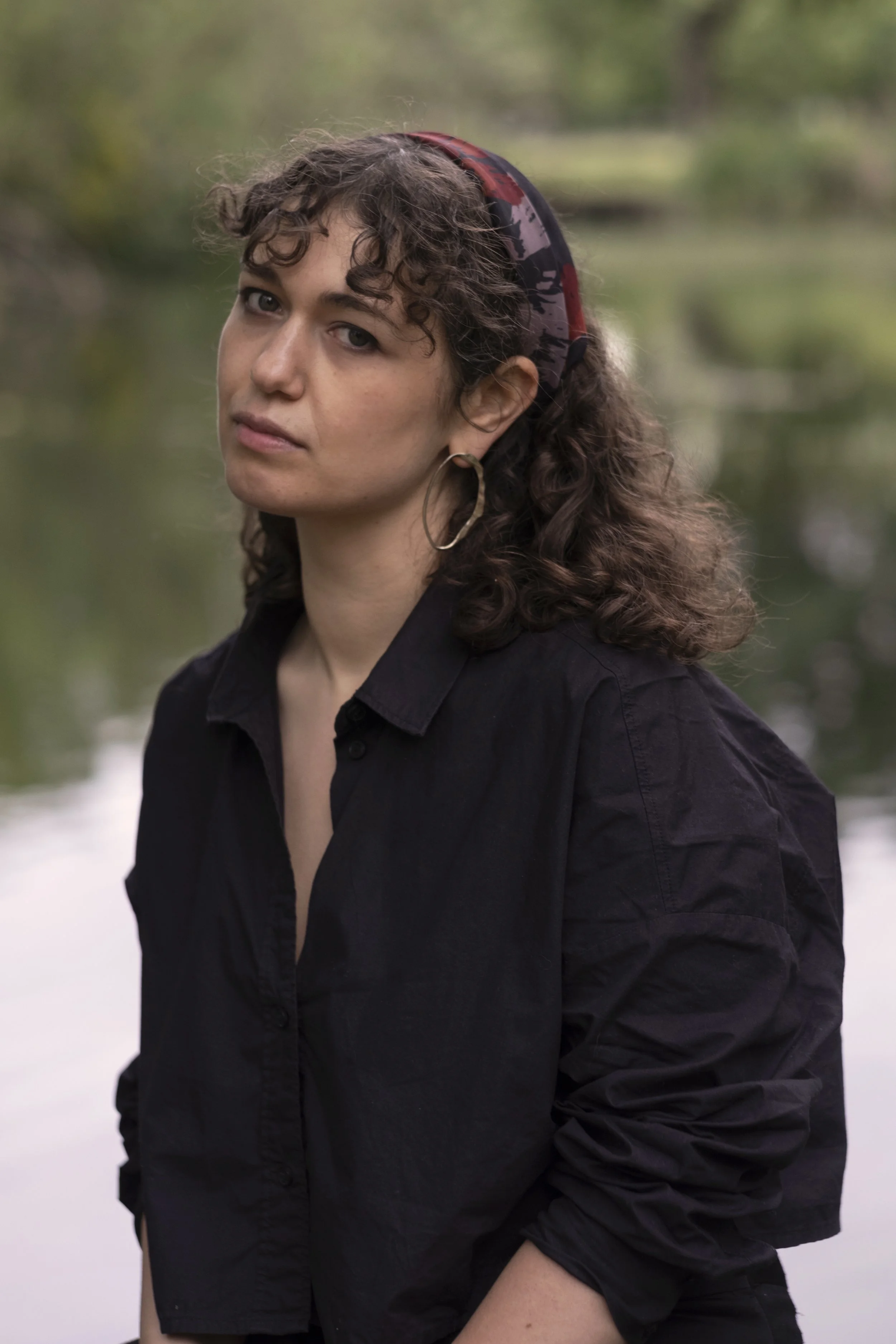 Portrait of a woman with curly hair wearing a headband, large hoop earrings, and a black shirt, standing outdoors near a body of water with blurred green trees in the background.