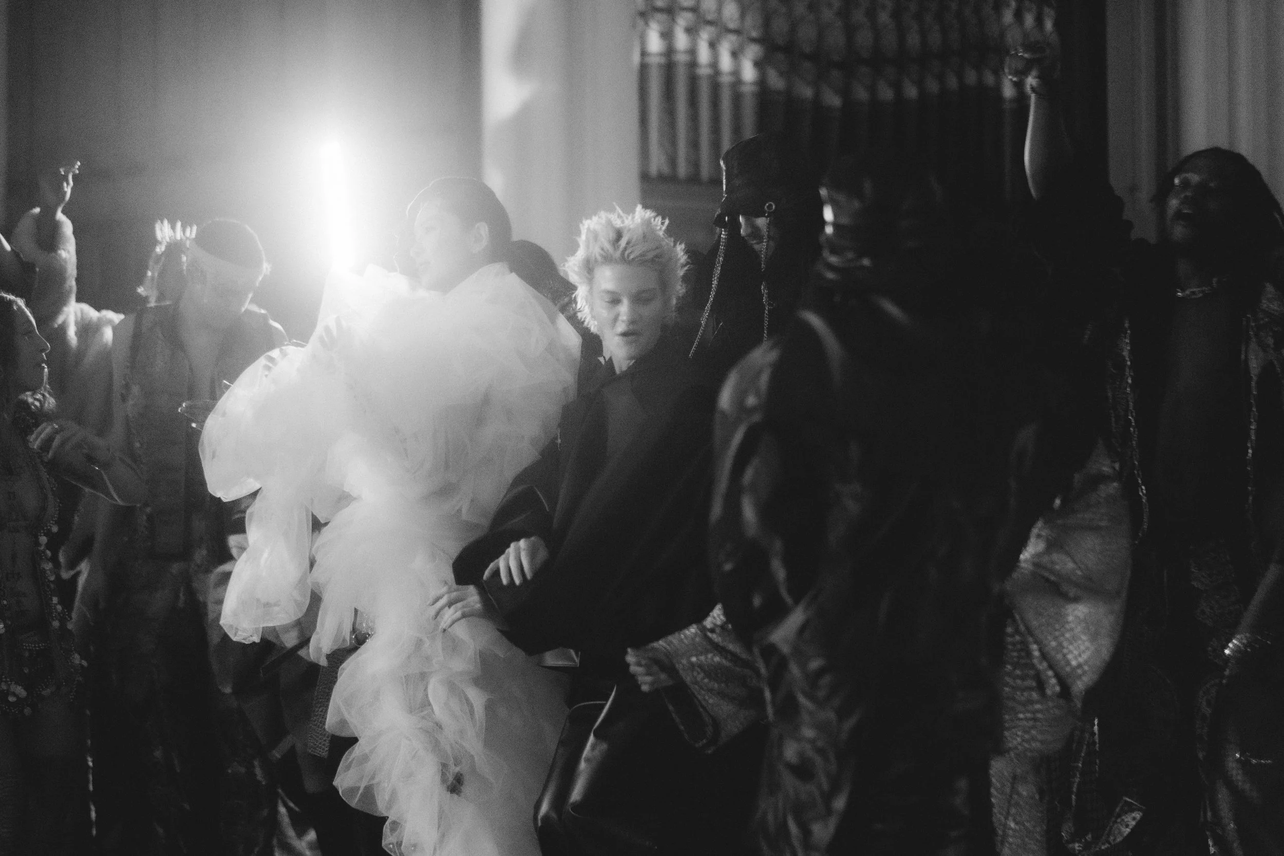 Black-and-white photo of a group of diverse people sitting and standing in a dimly lit room, some laughing and engaging with each other, with a strong light source creating a glow on the left side.