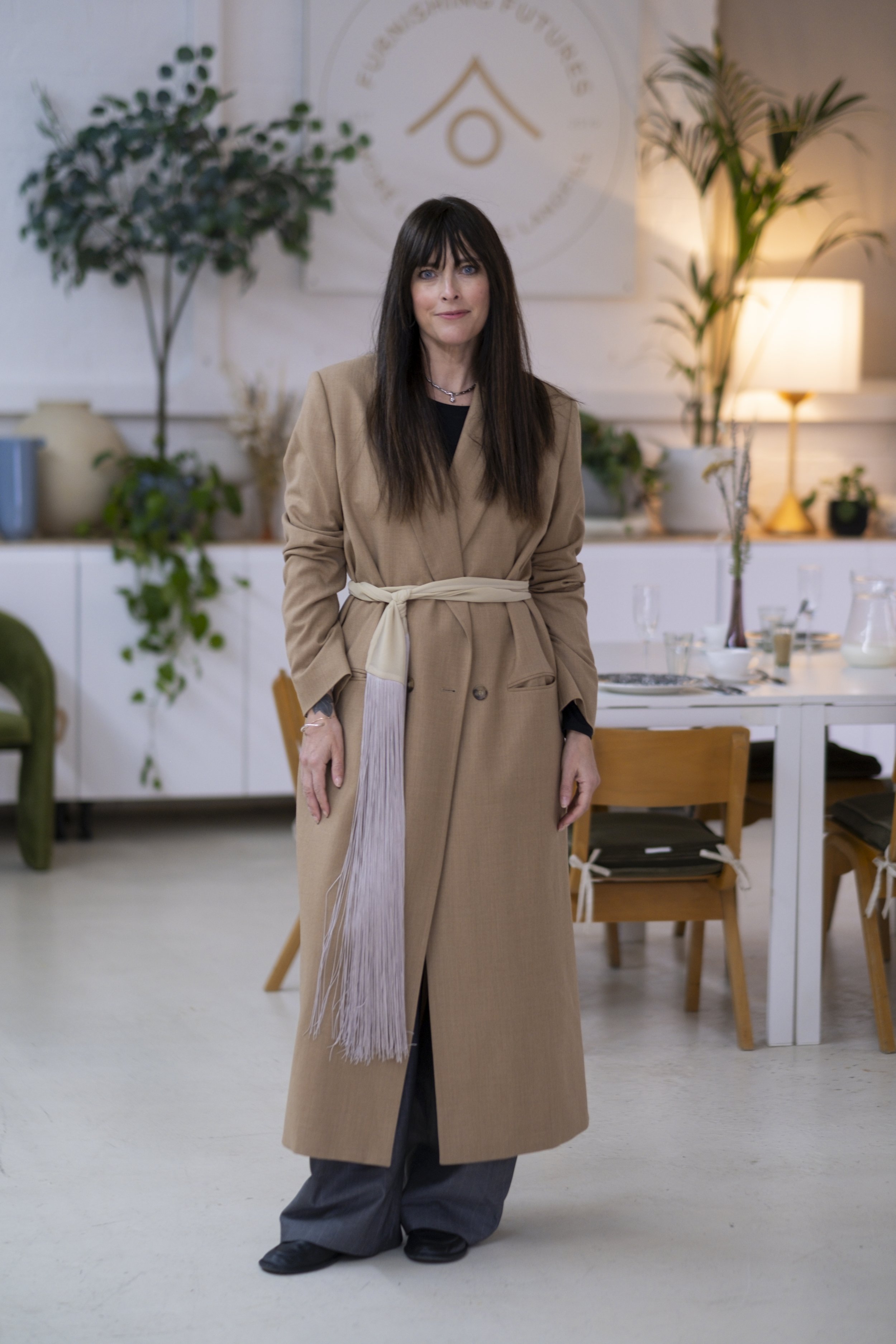 A woman with dark brown hair and bangs, wearing a tan trench coat with a white sash, black shirt, and dark pants, stands in a well-lit room with white walls, potted plants, and a table with empty glasses and a tall vase with dried flowers.