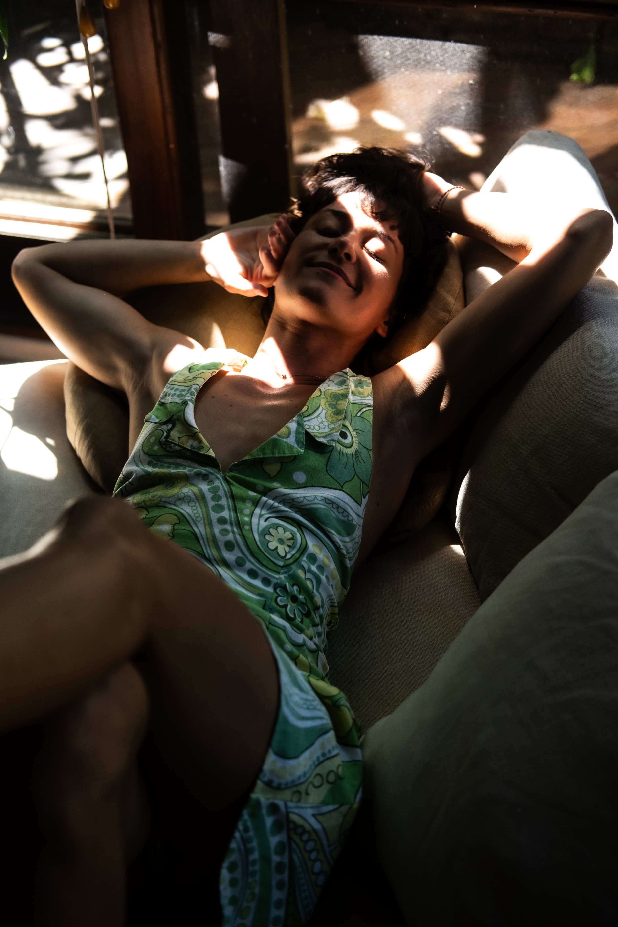 Young woman lying on a sofa with her eyes closed, smiling, and relaxing in sunlight, wearing a patterned green and yellow dress.