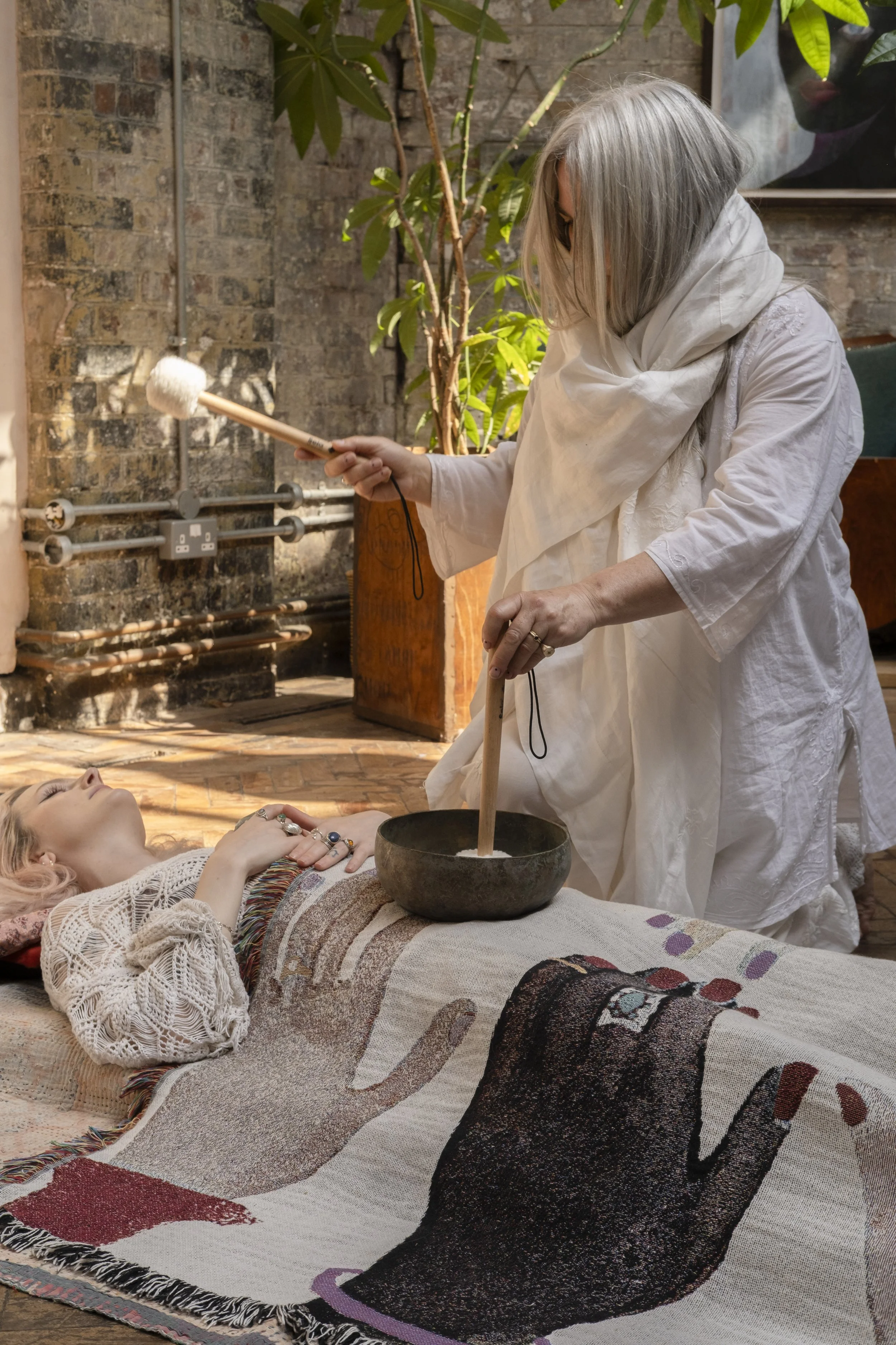 A woman with long gray hair dressed in white holds a long wooden stick with a white padded ball at the end, performing a healing or spiritual ritual over a reclining woman with curly hair, tattoos, and jewelry, lying on a large, colorful patterned rug. The setting is an indoor space with a brick wall, potted plants, and artwork.