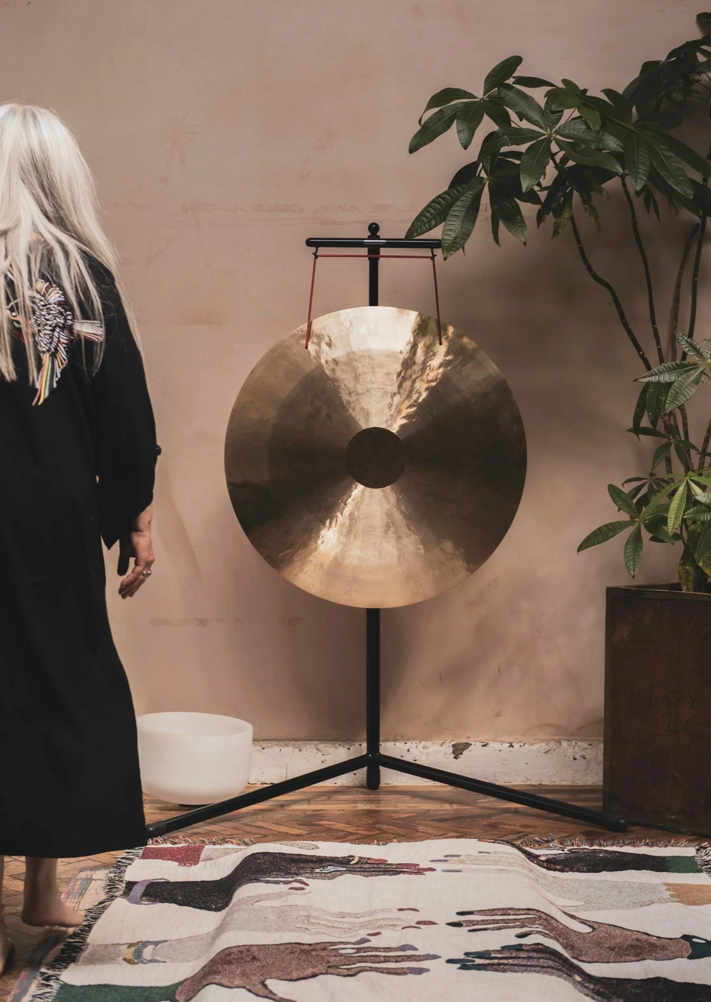 A room with a large metallic gong on a black stand, a green leafy plant in a pot, a colorful rug with abstract patterns, a white bowl, and a woman with long blonde hair wearing a black dress with colorful embroidery on the sleeve.