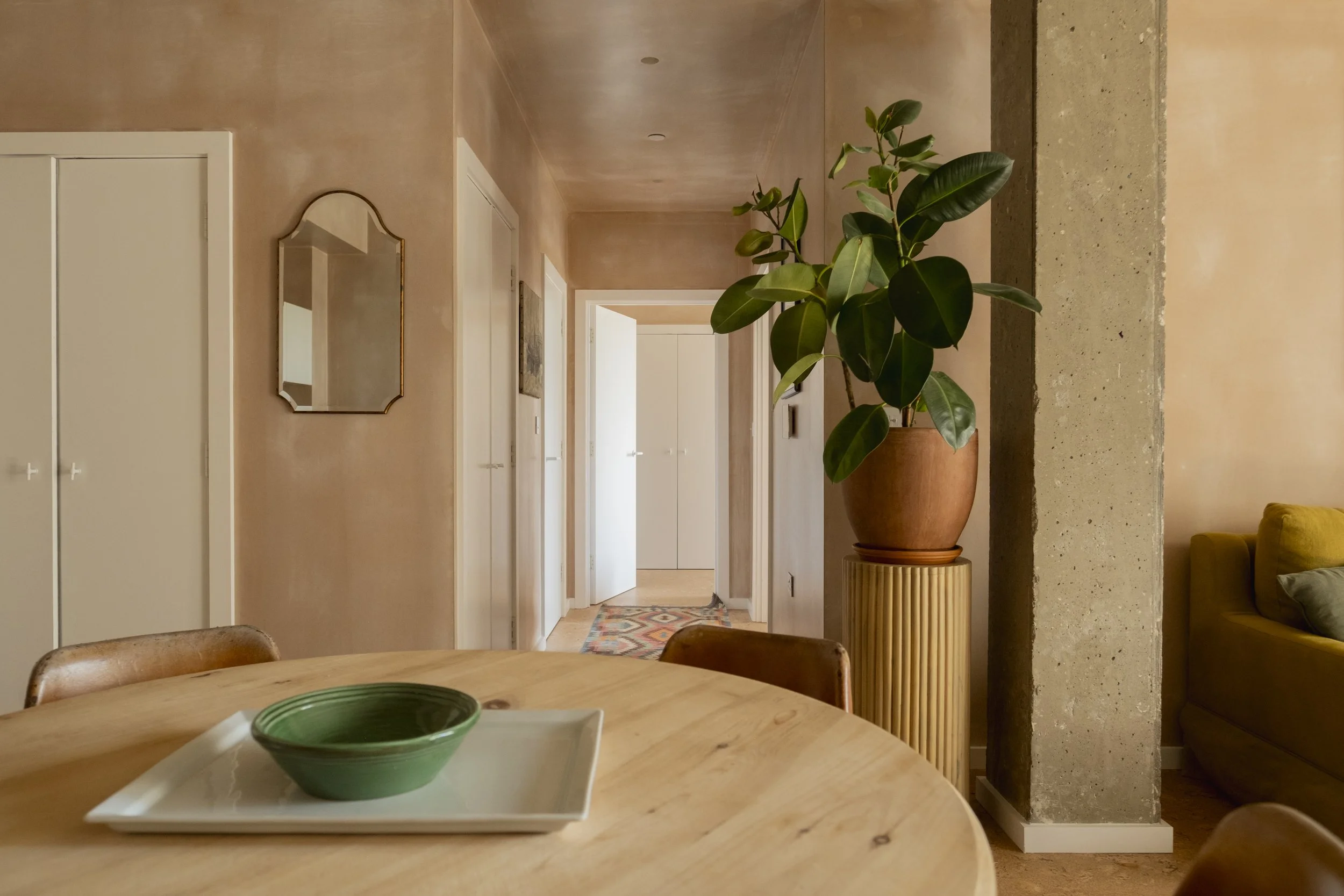 Interior view of a hallway seen from a dining table, with a potted plant on a stand and a mirror on the wall. The hallway has beige walls and white doors.