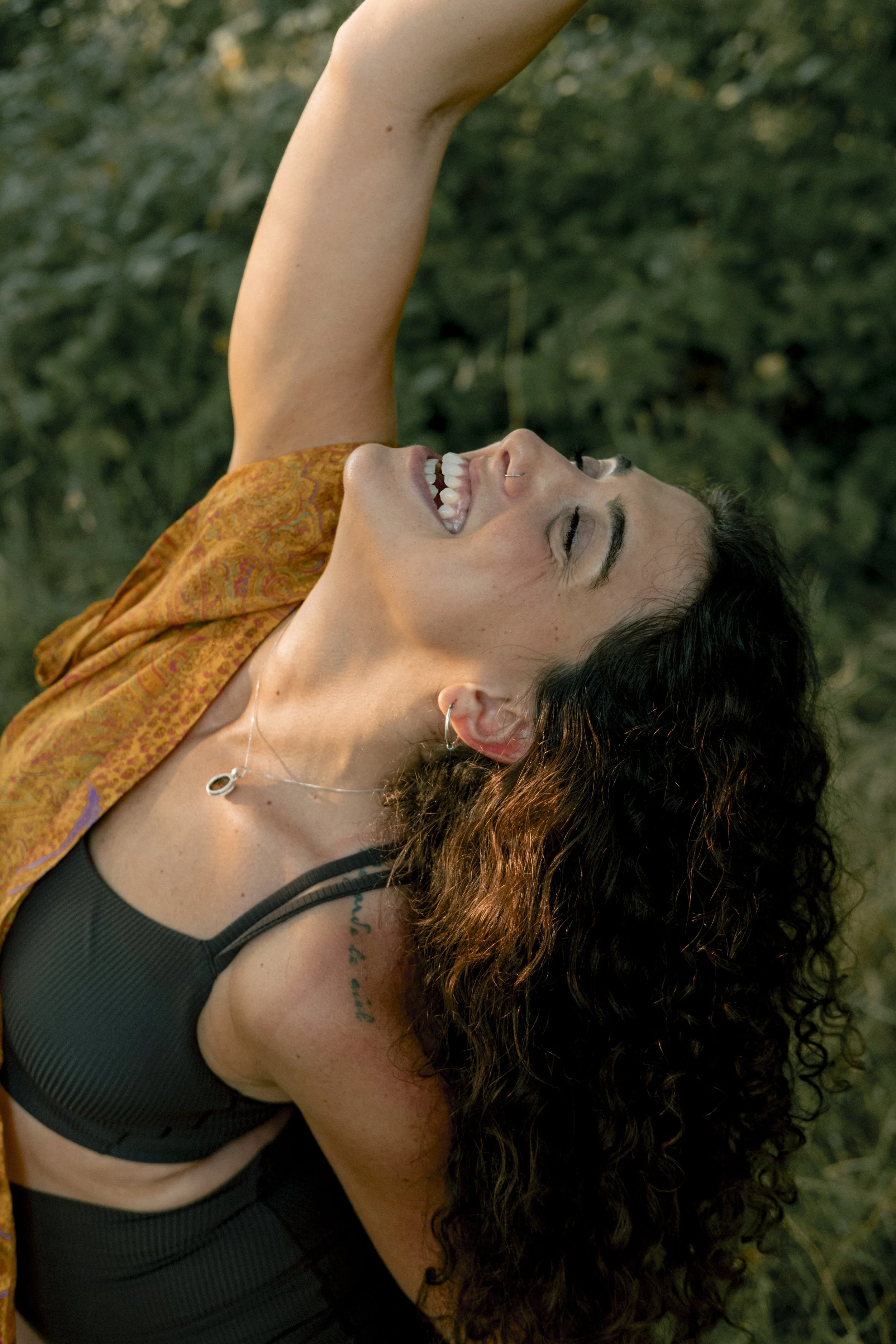 A woman with curly dark hair, wearing a black sports bra and some jewelry, is smiling with her eyes closed and appears to be outdoors in a green, leafy environment.