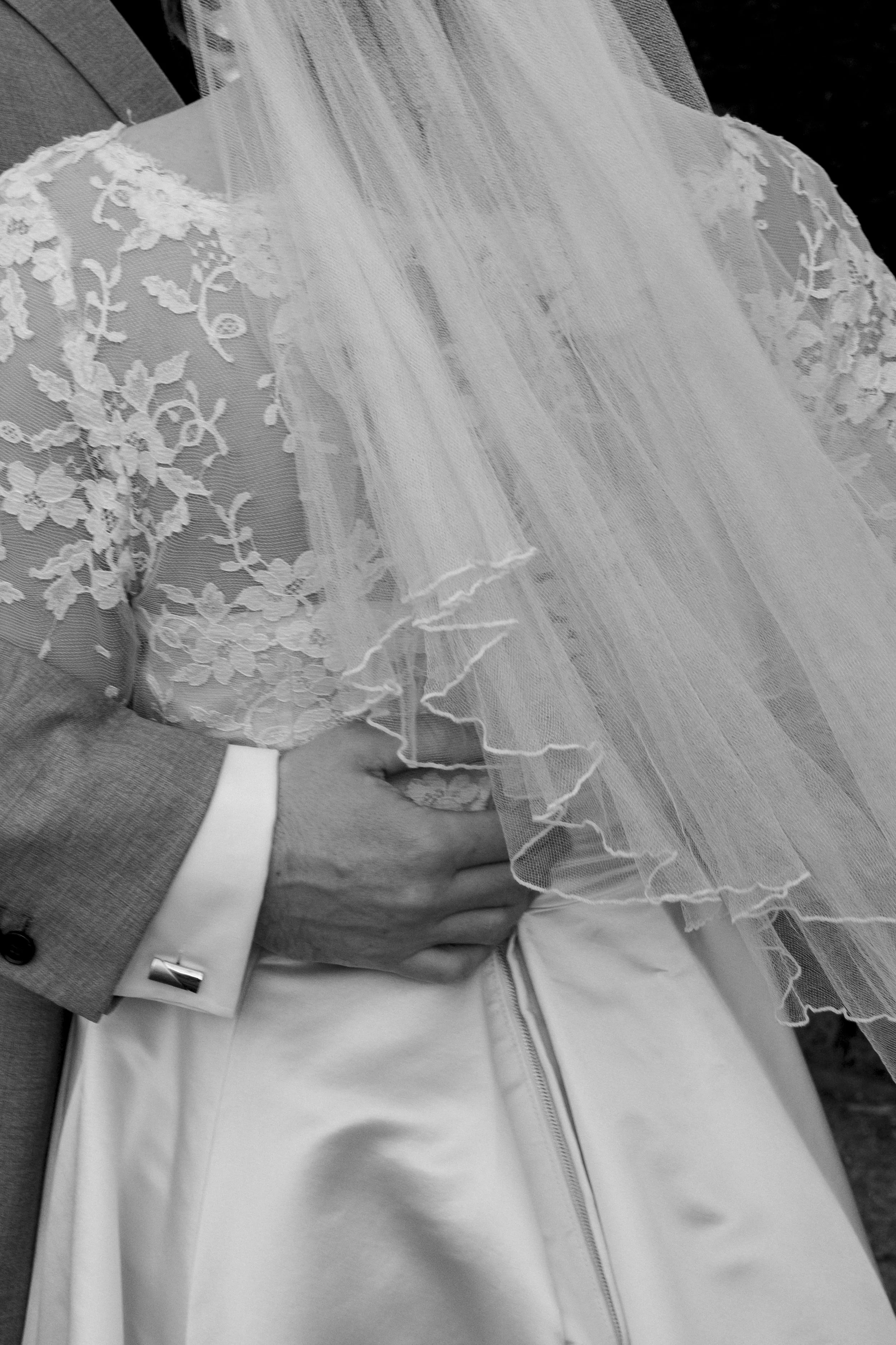 Close-up black and white photo of a wedding dress with lace and tulle fabric, worn by a seated bride outside. A man in a suit is sitting beside her, with his hand on her arm.