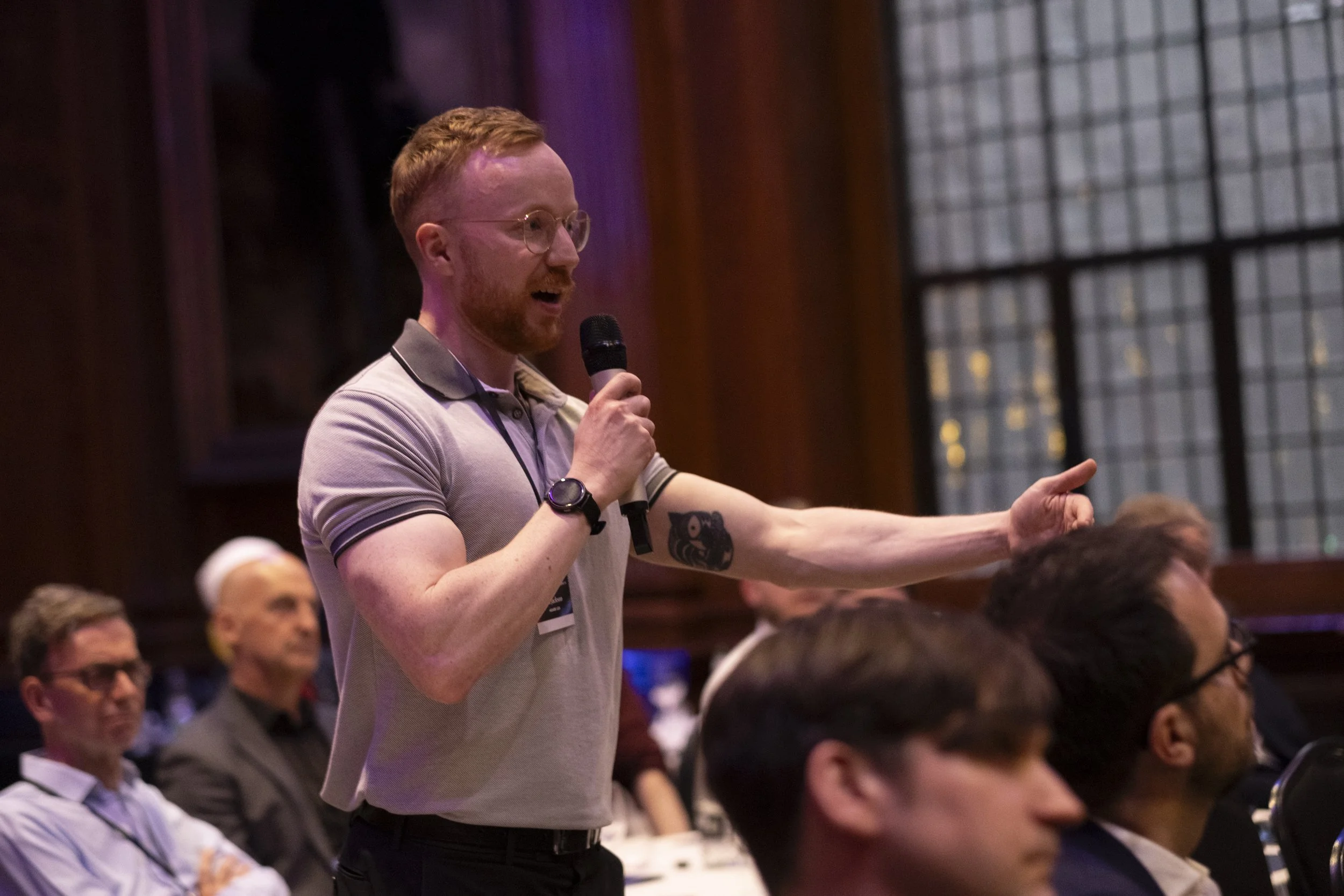 A man with red hair, glasses, and a beard speaking into a microphone during a conference or event, with several people seated and listening in a dimly lit room.