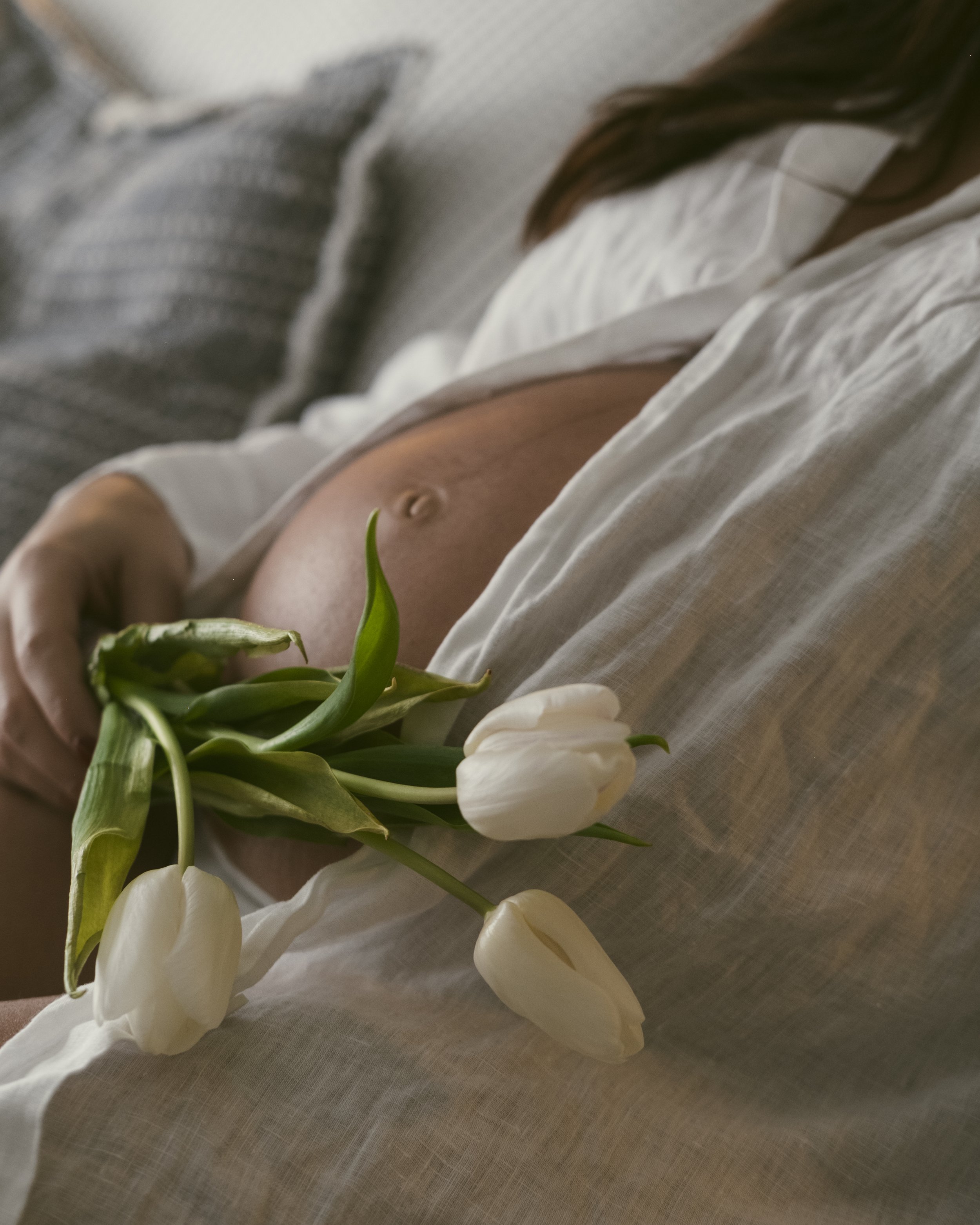 Close-up of a pregnant woman holding a bouquet of white tulips, wearing a white top and light gray cardigan, with the focus on her belly and flowers.