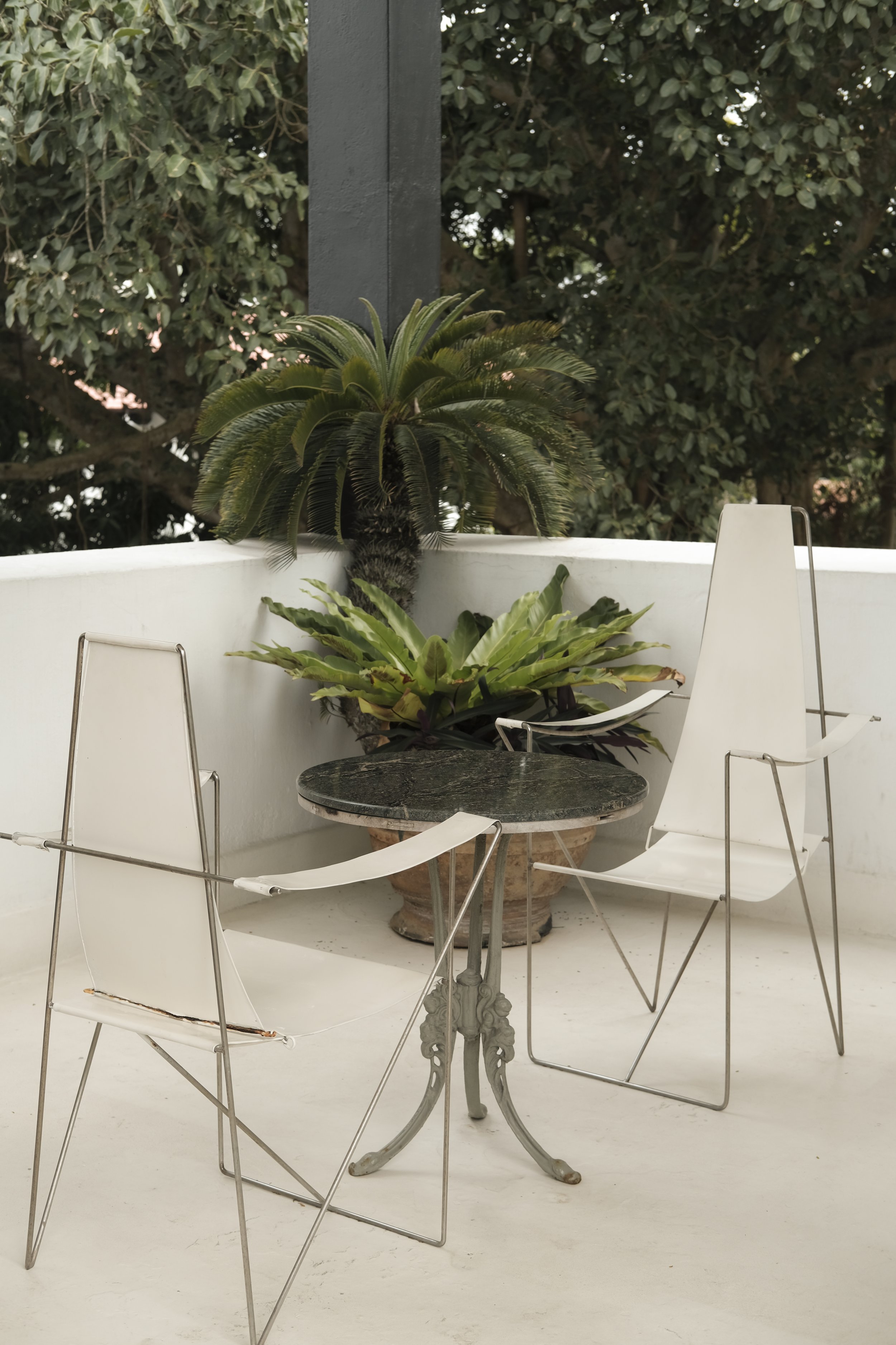 Outdoor patio with white chairs surrounding a small round black marble table under a white wall with planters containing green plants, and trees in the background.
