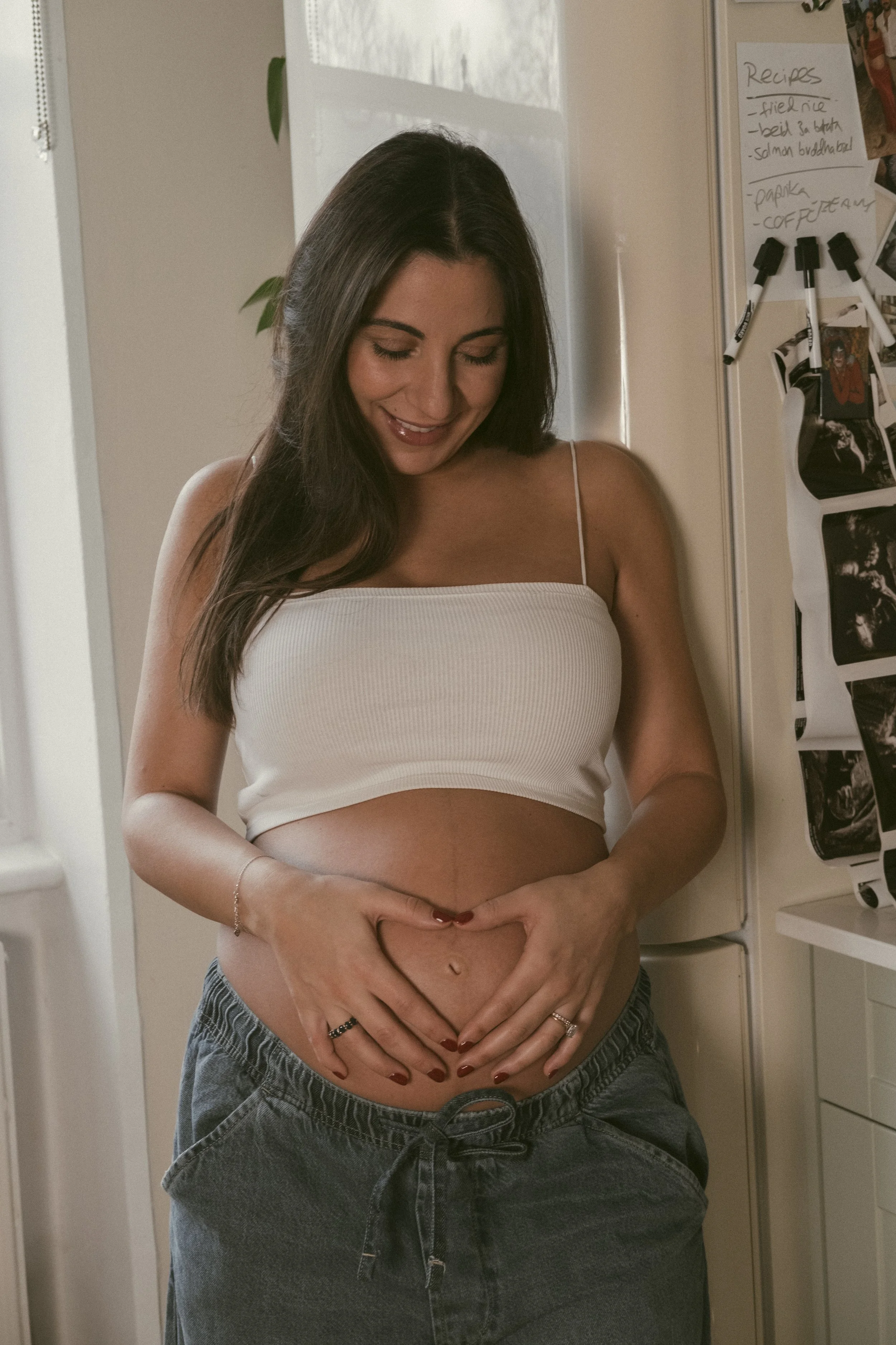A woman with long brown hair, wearing a white tank top, smiling with her eyes closed, standing in front of a refrigerator in a kitchen. She has her hands in a heart shape over her pregnant stomach.