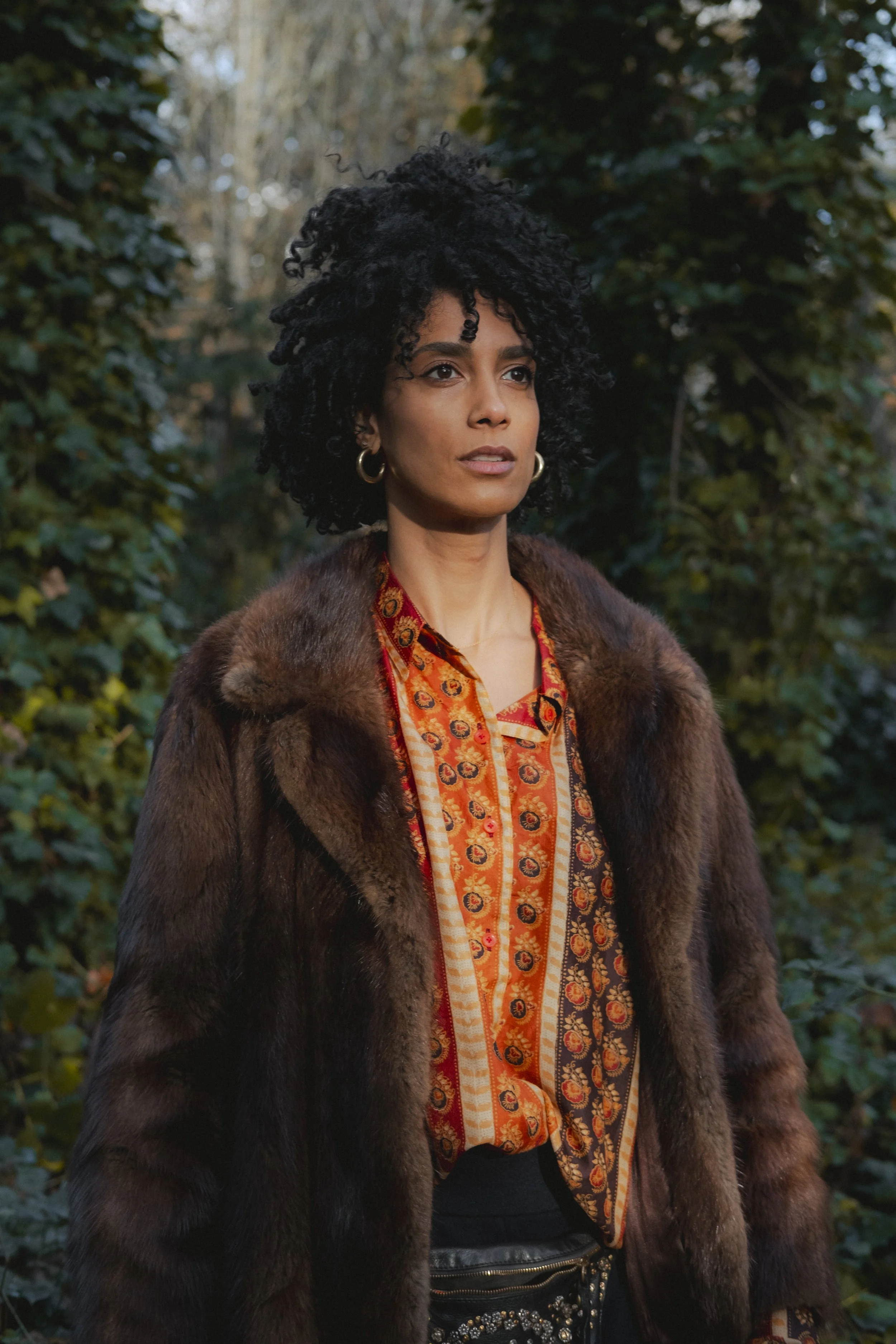 Woman with dark curly hair wearing gold hoop earrings and a luxurious fur coat standing outdoors with green foliage in the background.