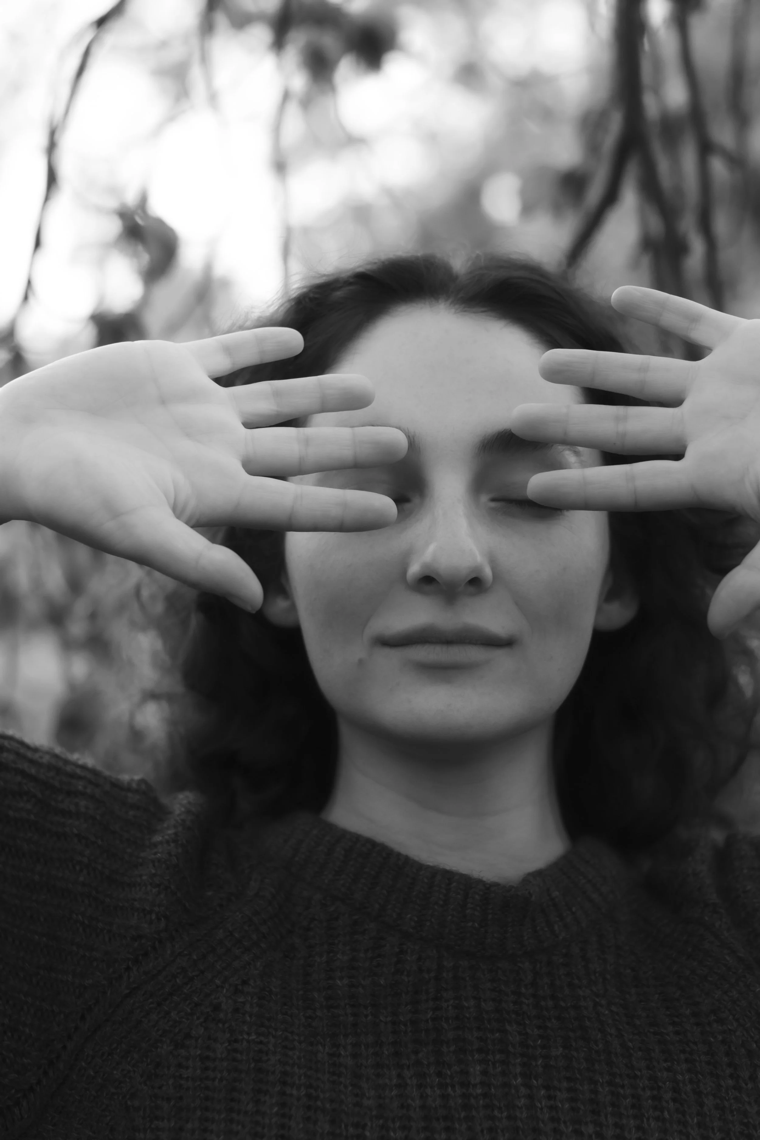 Black and white photo of a woman with dark hair, closing her eyes with a calm expression, hands framing her face, background of tree branches and leaves.