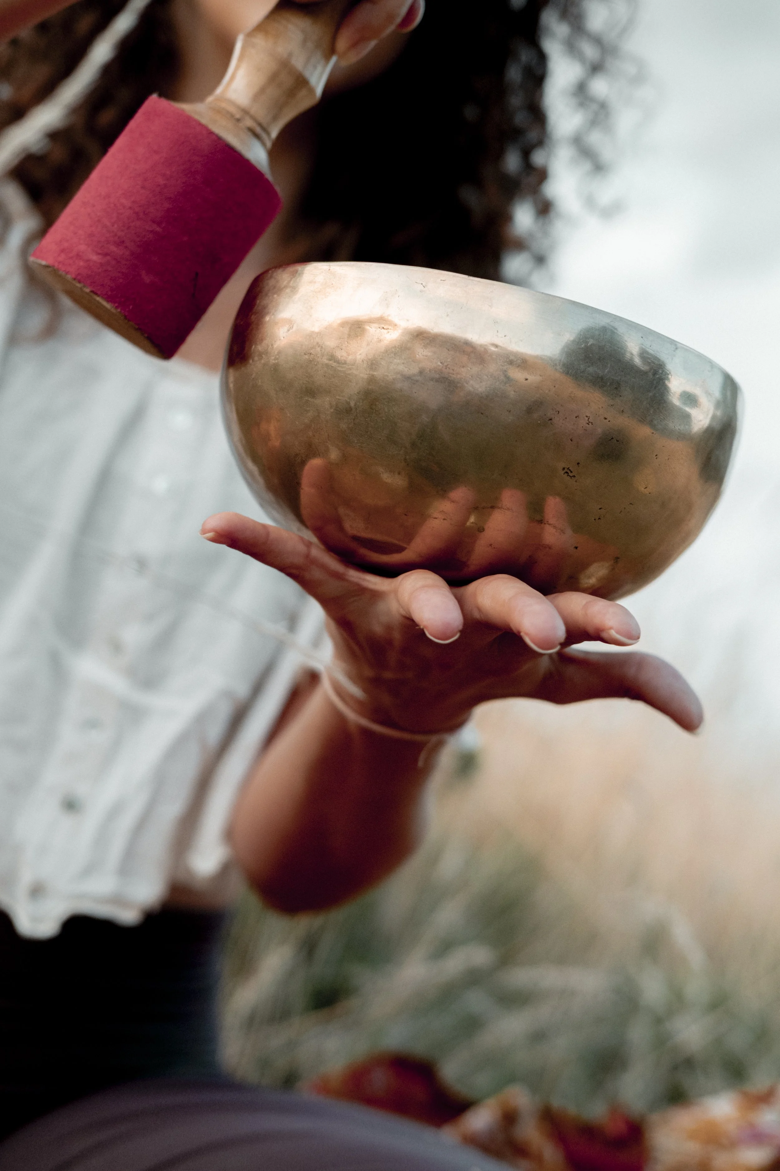 A person holding a metallic singing bowl with their fingers, with a red wristband on their wrist and wearing a white shirt with buttons.