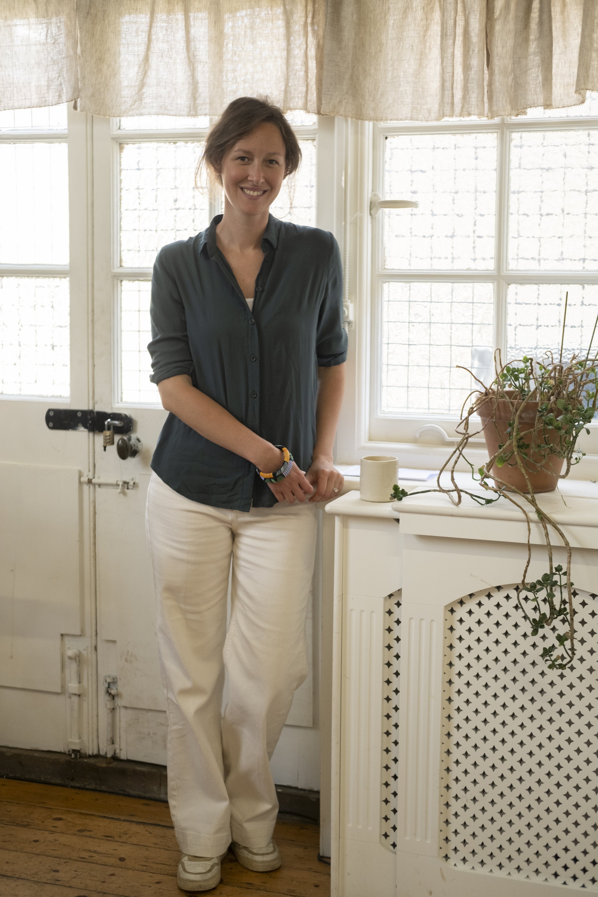 A woman with brown hair, wearing a black button-up shirt and white pants, standing indoors near a window with beige curtains. There is a potted plant and some cups on a white radiator cover.