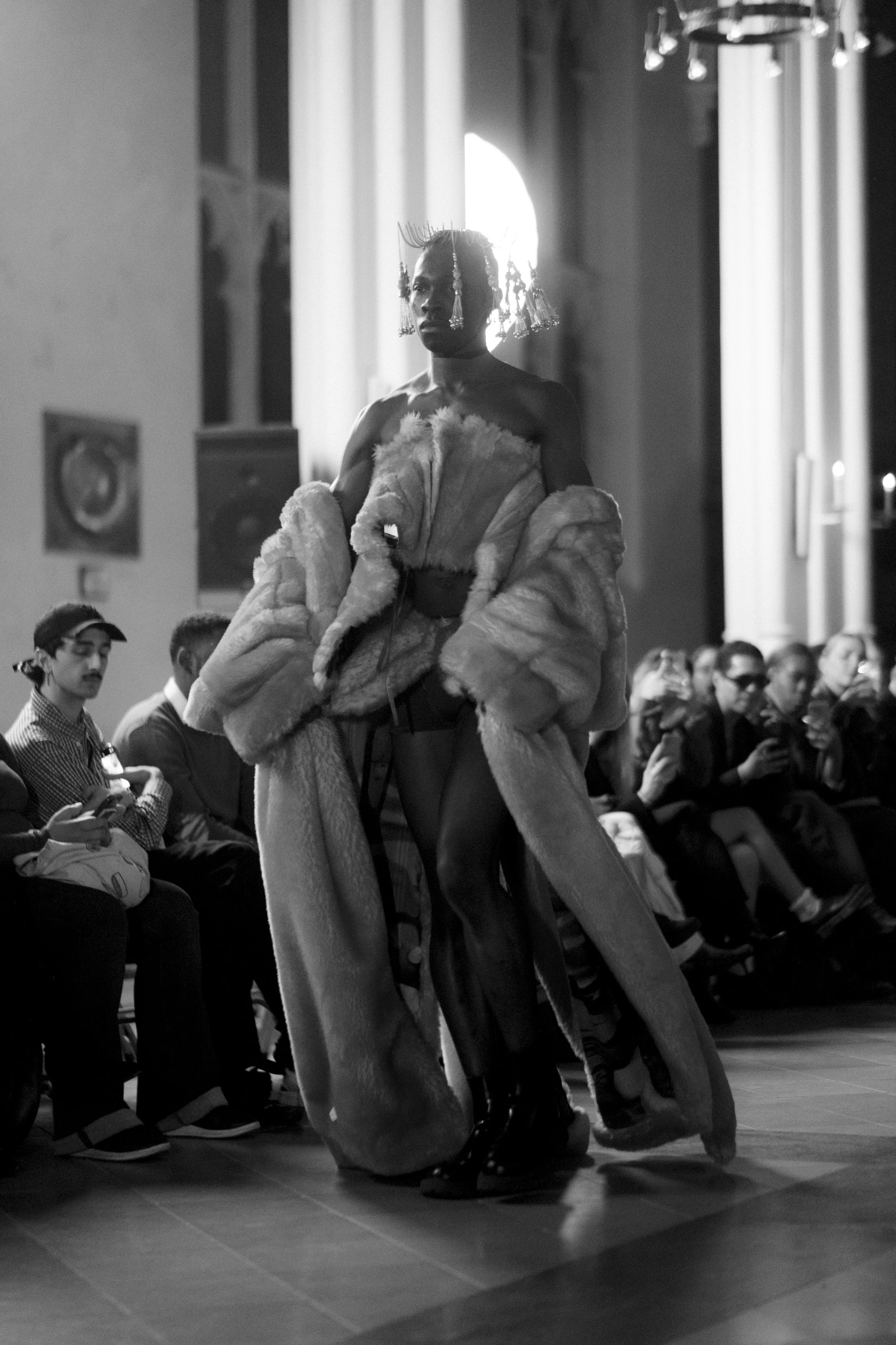 A black-and-white photograph captures a person walking down a runway at a fashion show, wearing a long fur coat and statement jewelry, with an audience seated on either side watching.