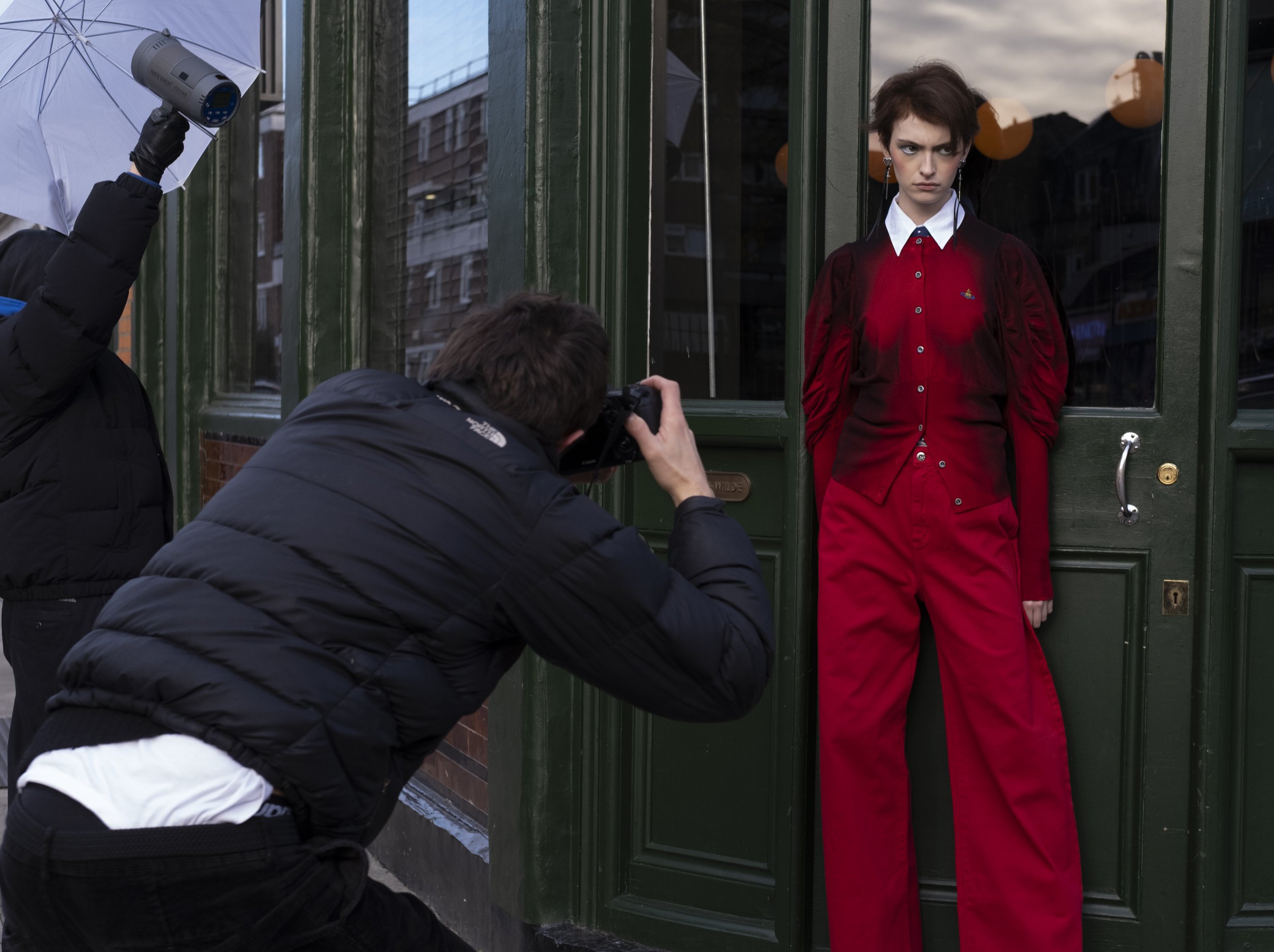 Two photographers are taking pictures of a woman standing against a green door. The woman has short dark hair, is wearing a red jacket over a white shirt, and has a serious expression.