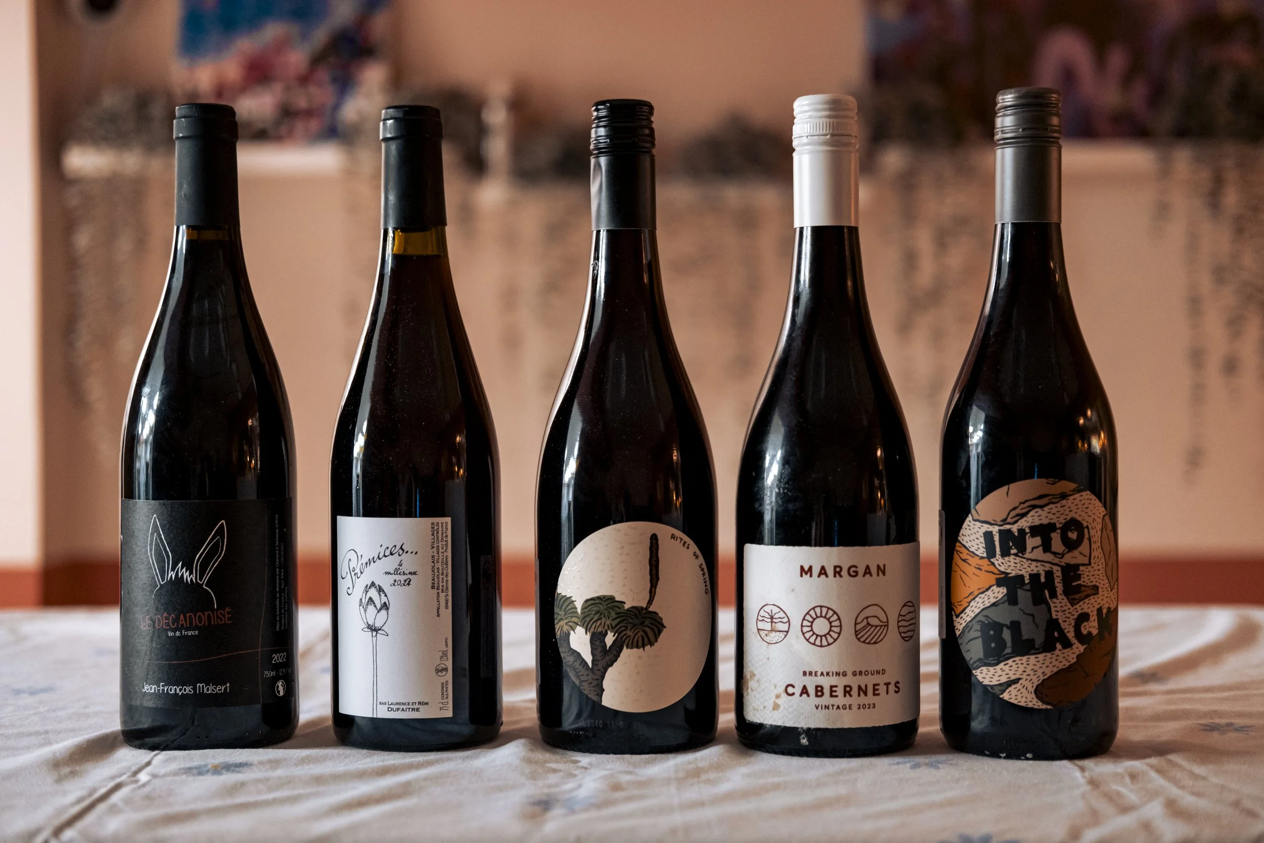 Five bottles of wine lined up on a table with a blurred background, each with distinct labels and caps.