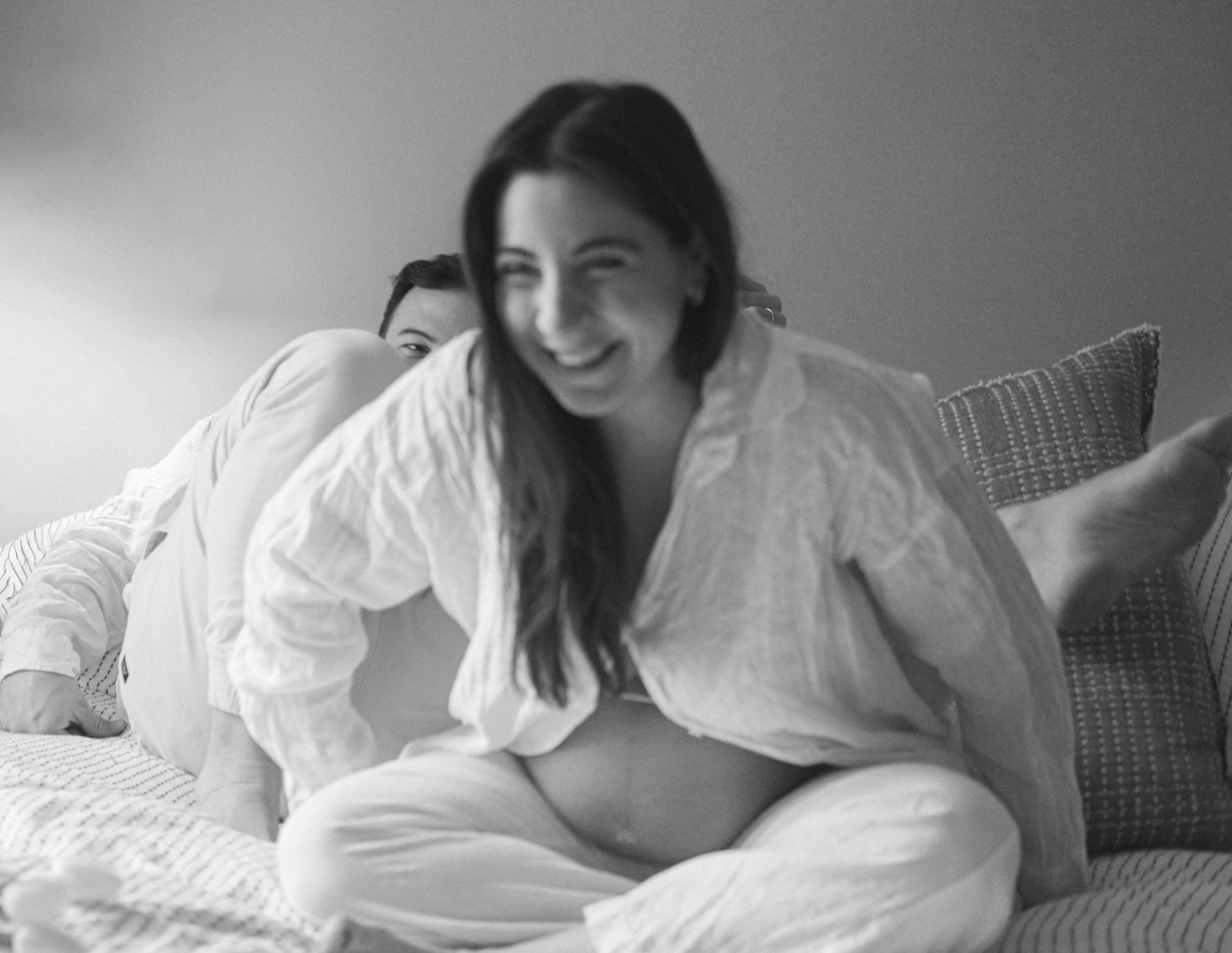 Two women lying on a bed, one with dark shoulder-length hair is smiling, while the other is partly hidden behind her. The image is in black and white.