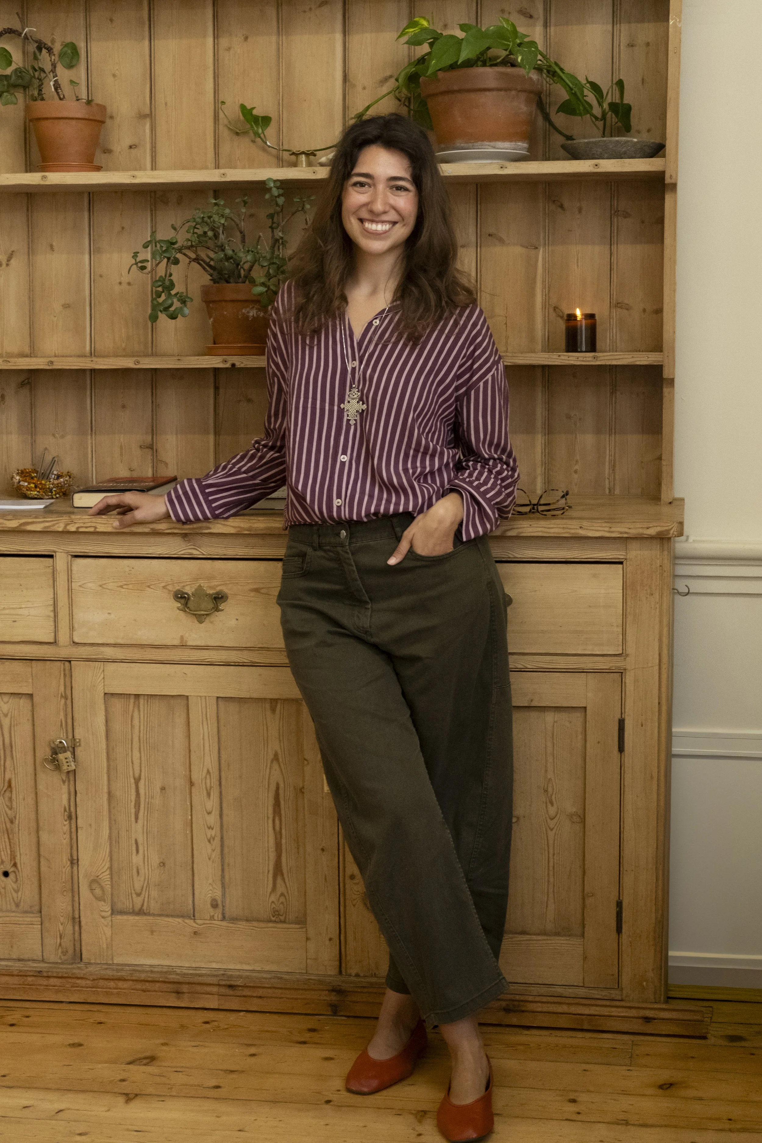 A woman with shoulder-length dark hair, wearing a striped purple blouse and black pants, standing in front of a wooden shelf with plants and a lit candle, smiling.