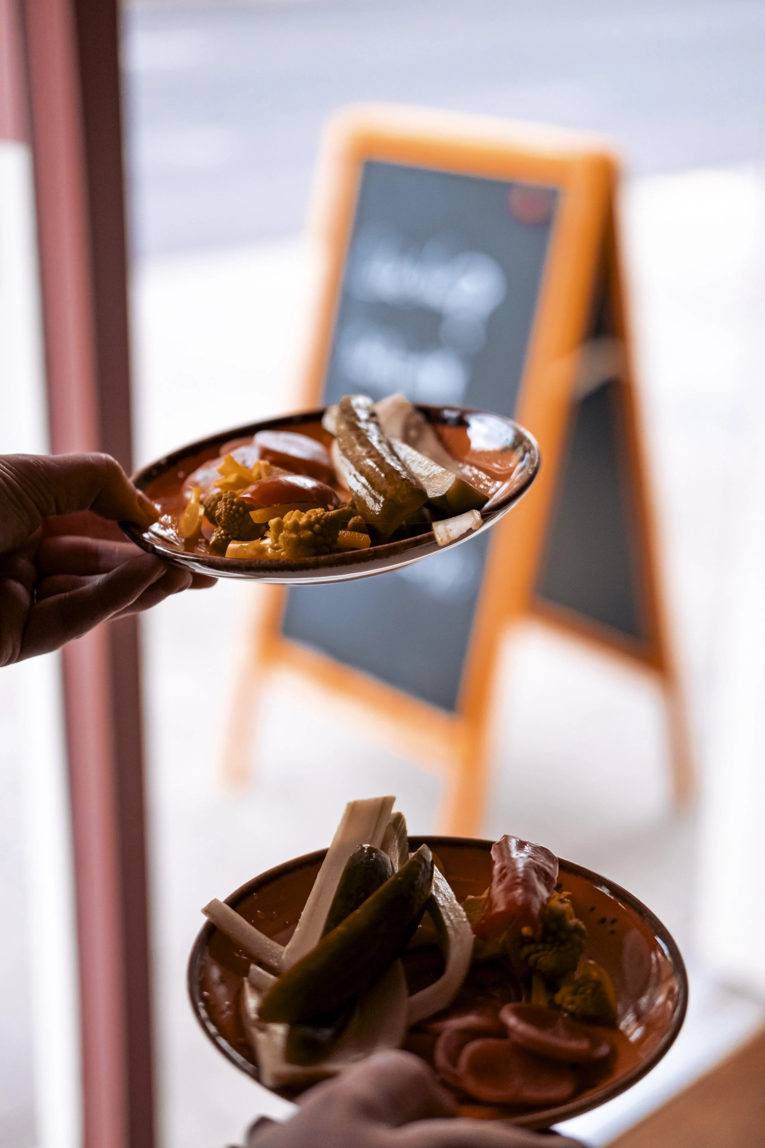 Close-up of a hand holding a small bowl of mixed pickled vegetables, with a blurry menu or sign board in the background.