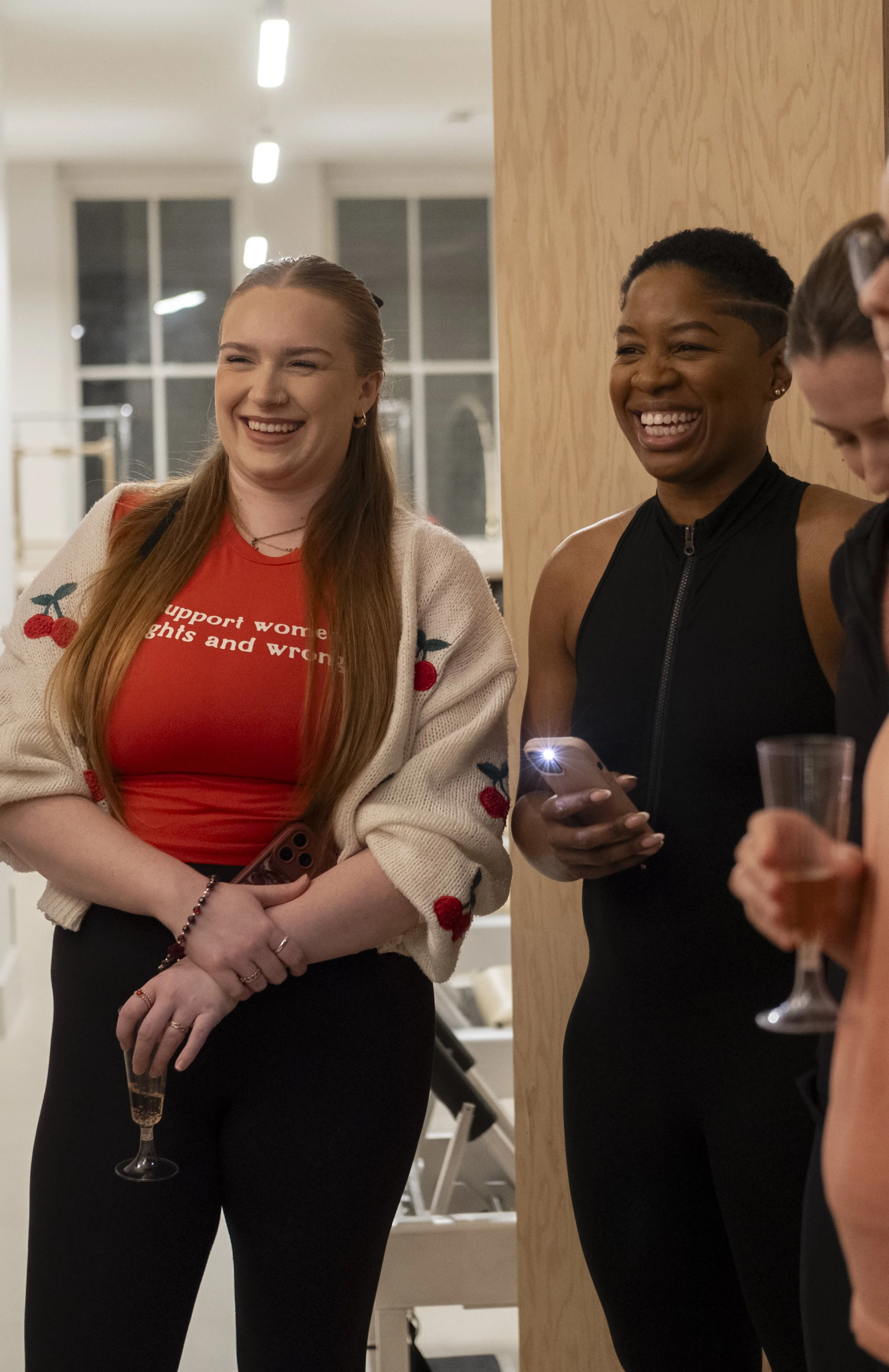 Two women smiling and laughing at a social gathering, one holding a glass of champagne and the other holding a smartphone.