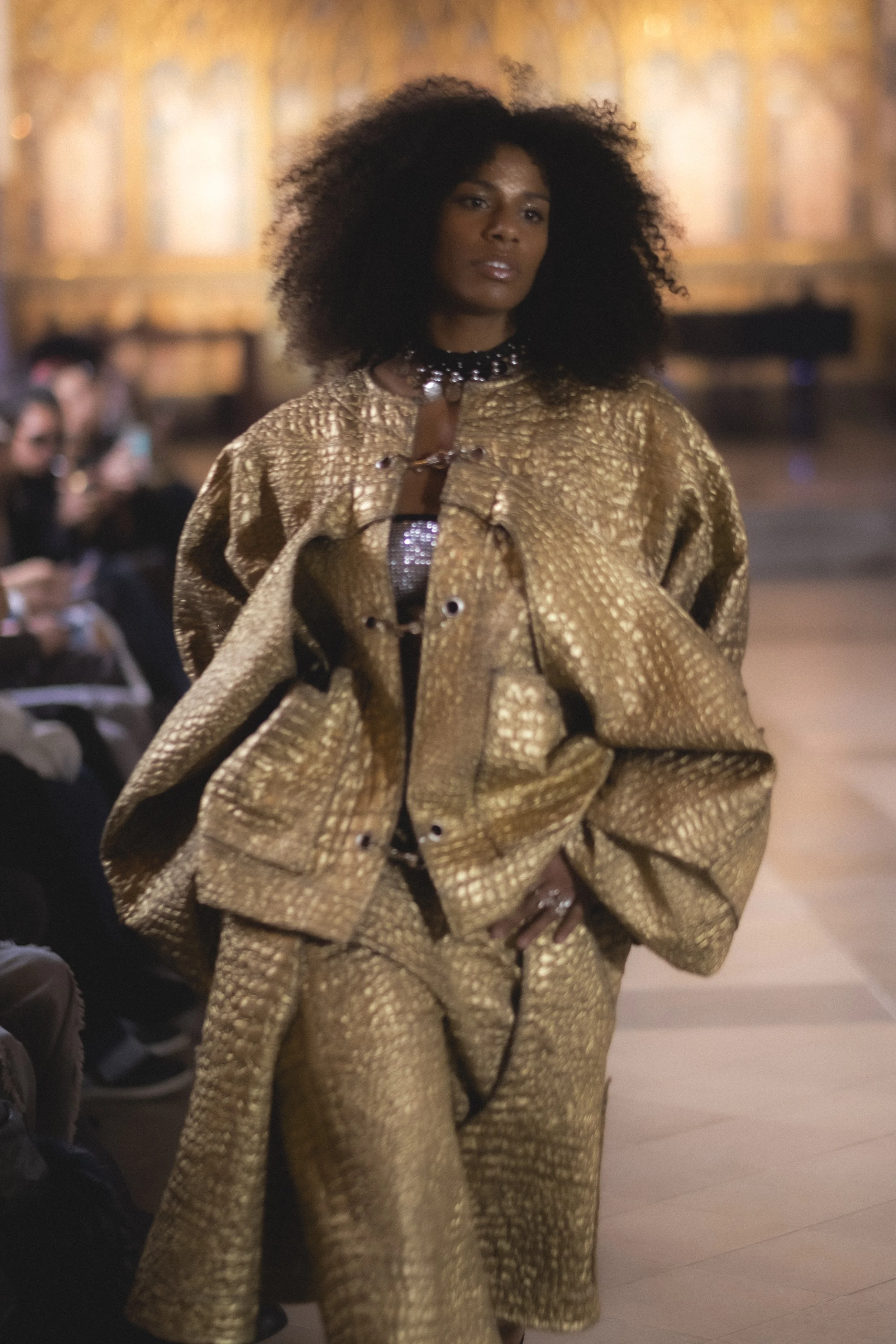 A woman with curly black hair walking on a runway, wearing a gold textured coat with a matching dress underneath, and accessorized with a pearl choker necklace with a brooch, during a fashion show.