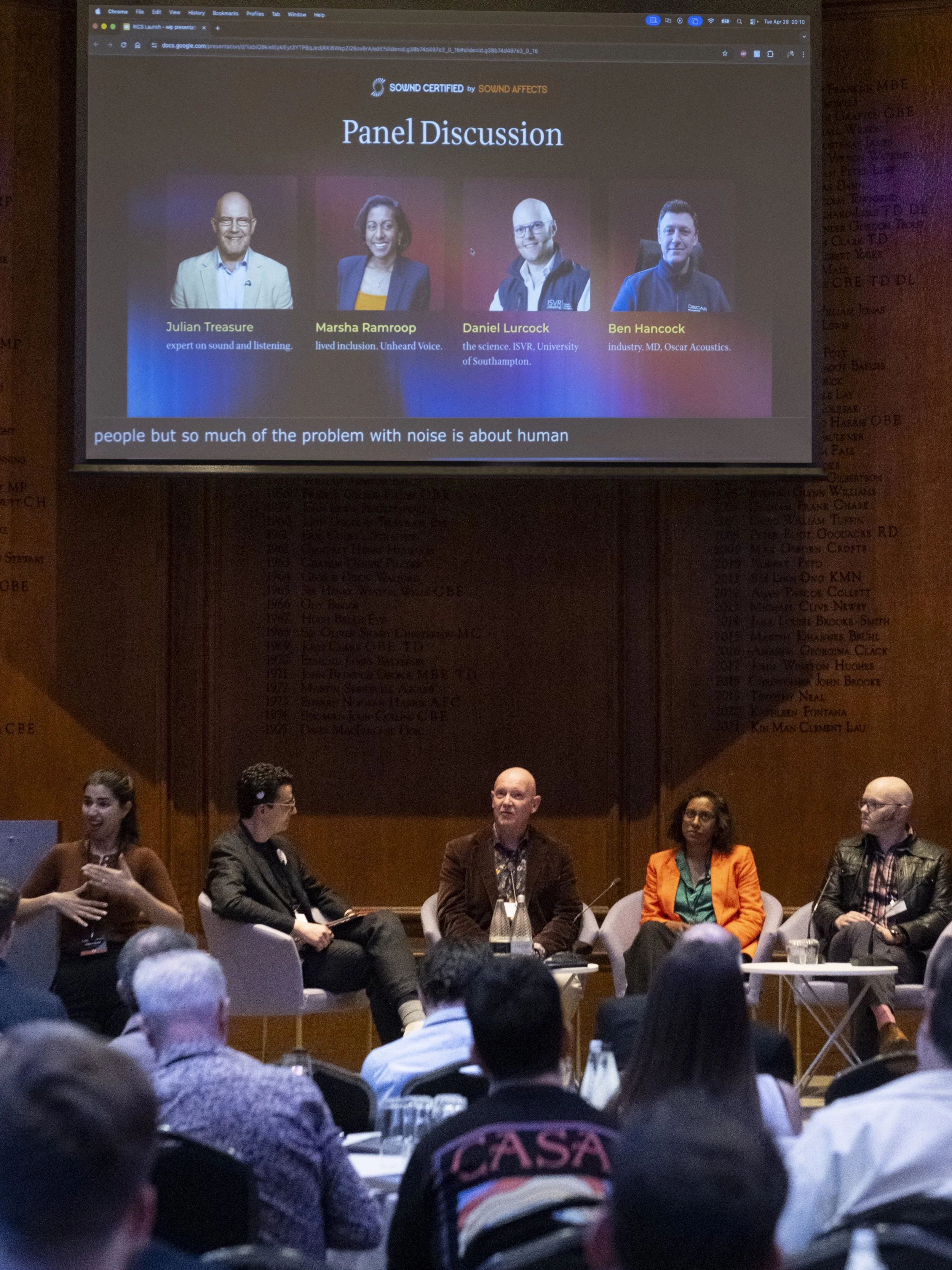 A panel discussion with four individuals seated on stage in front of an audience. A large screen behind them displays the panel title and individual names and titles.