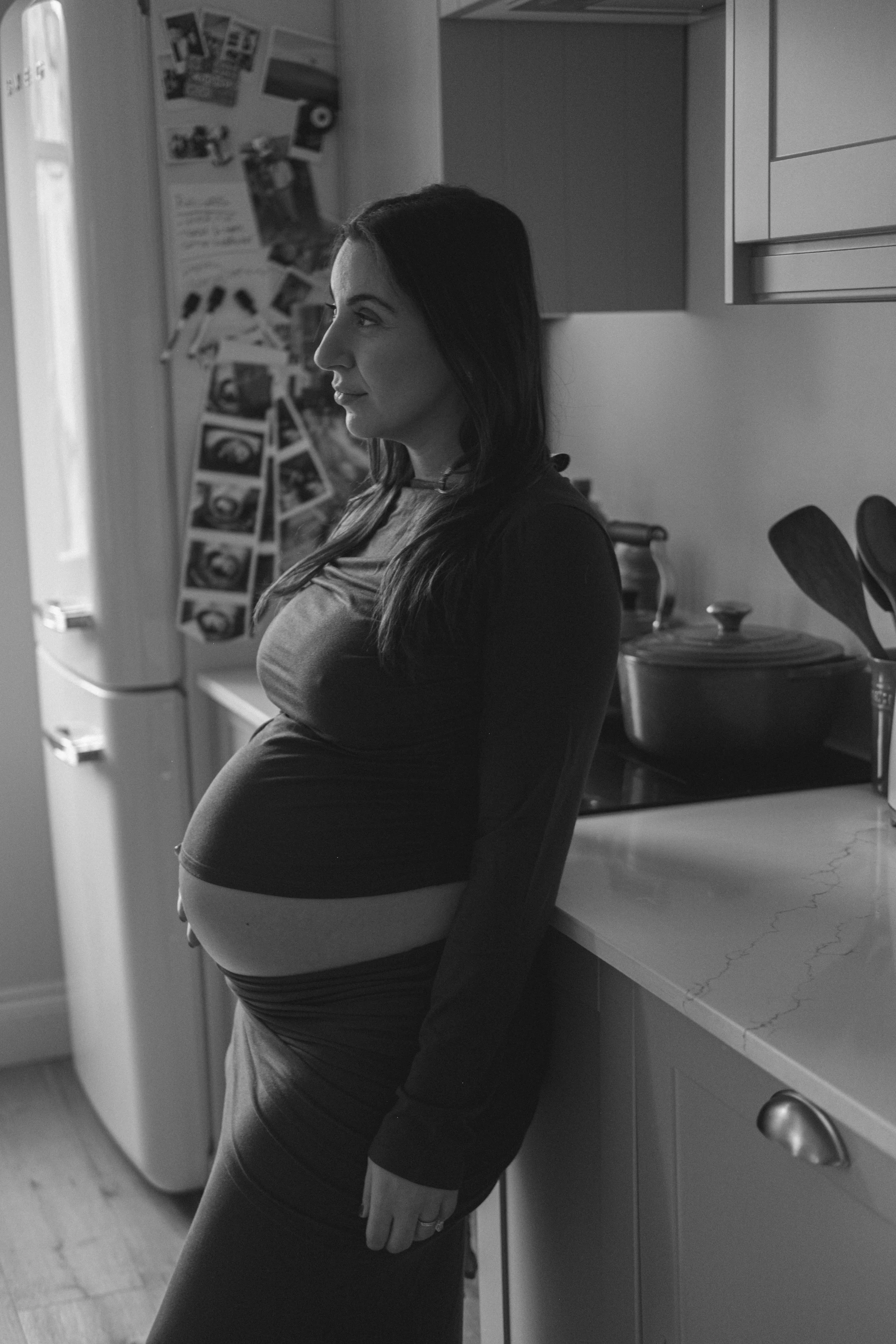 A woman with dark hair, wearing a long-sleeved shirt and leggings, stands in a kitchen with her back against a counter, looking to her left. The kitchen has a refrigerator with photos and notes, and various kitchen utensils and pots are visible.