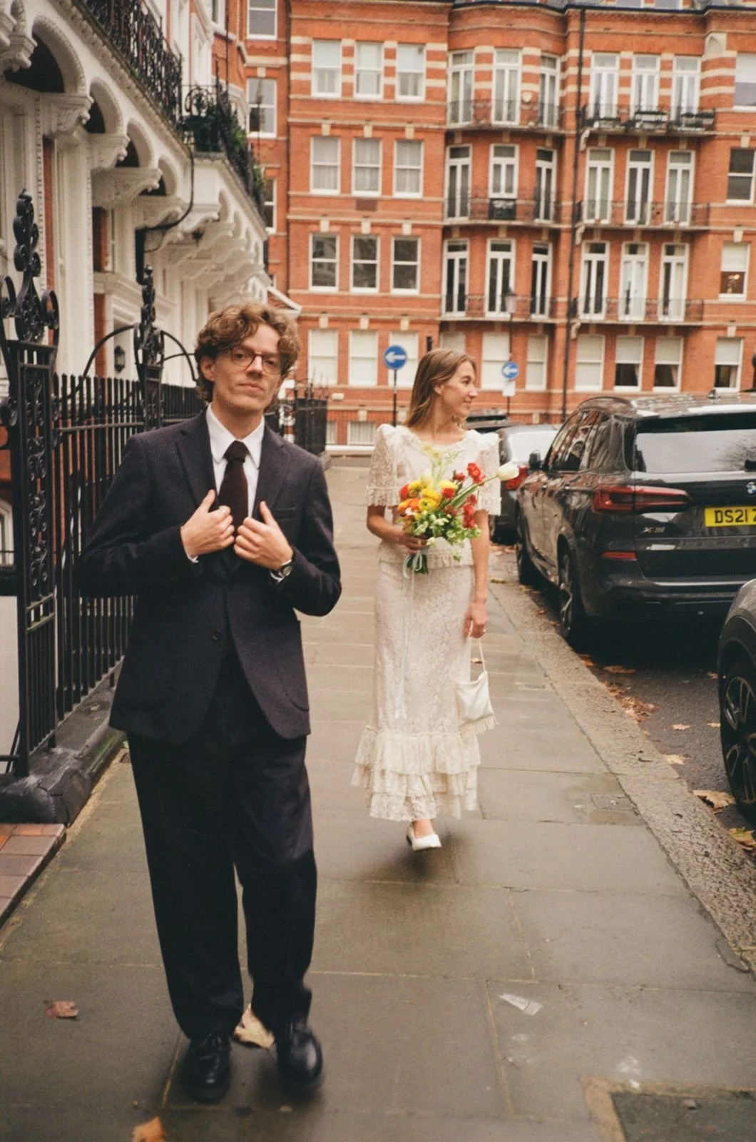 A man in a suit and tie stands on a city sidewalk, adjusting his jacket, with a woman in a vintage white lace dress holding a bouquet and small handbag walking behind him, in front of red-brick apartment buildings and parked cars.
