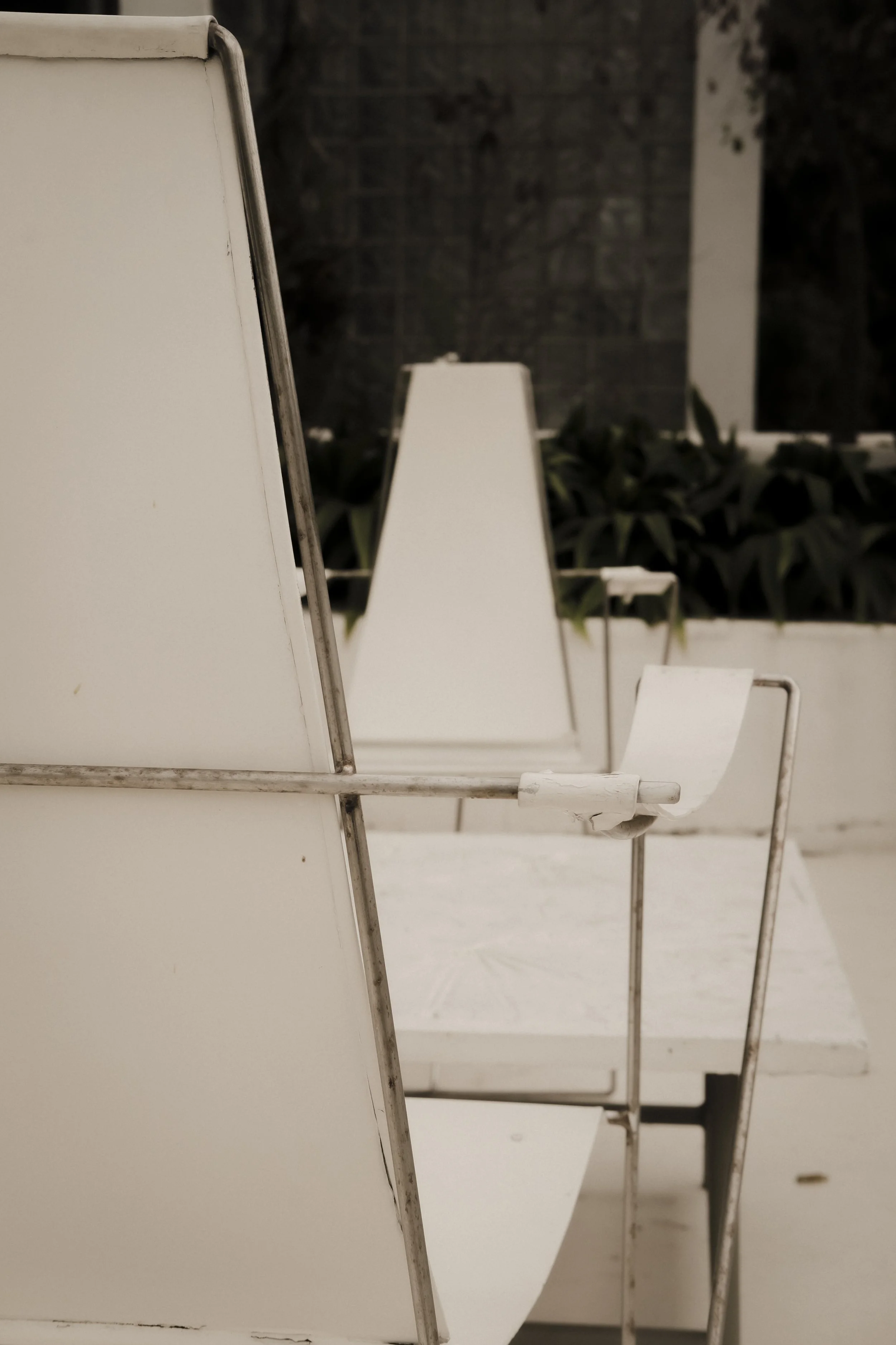 White metal chairs with white tabletops arranged in a row outdoors, with plants and a dark wall in the background.