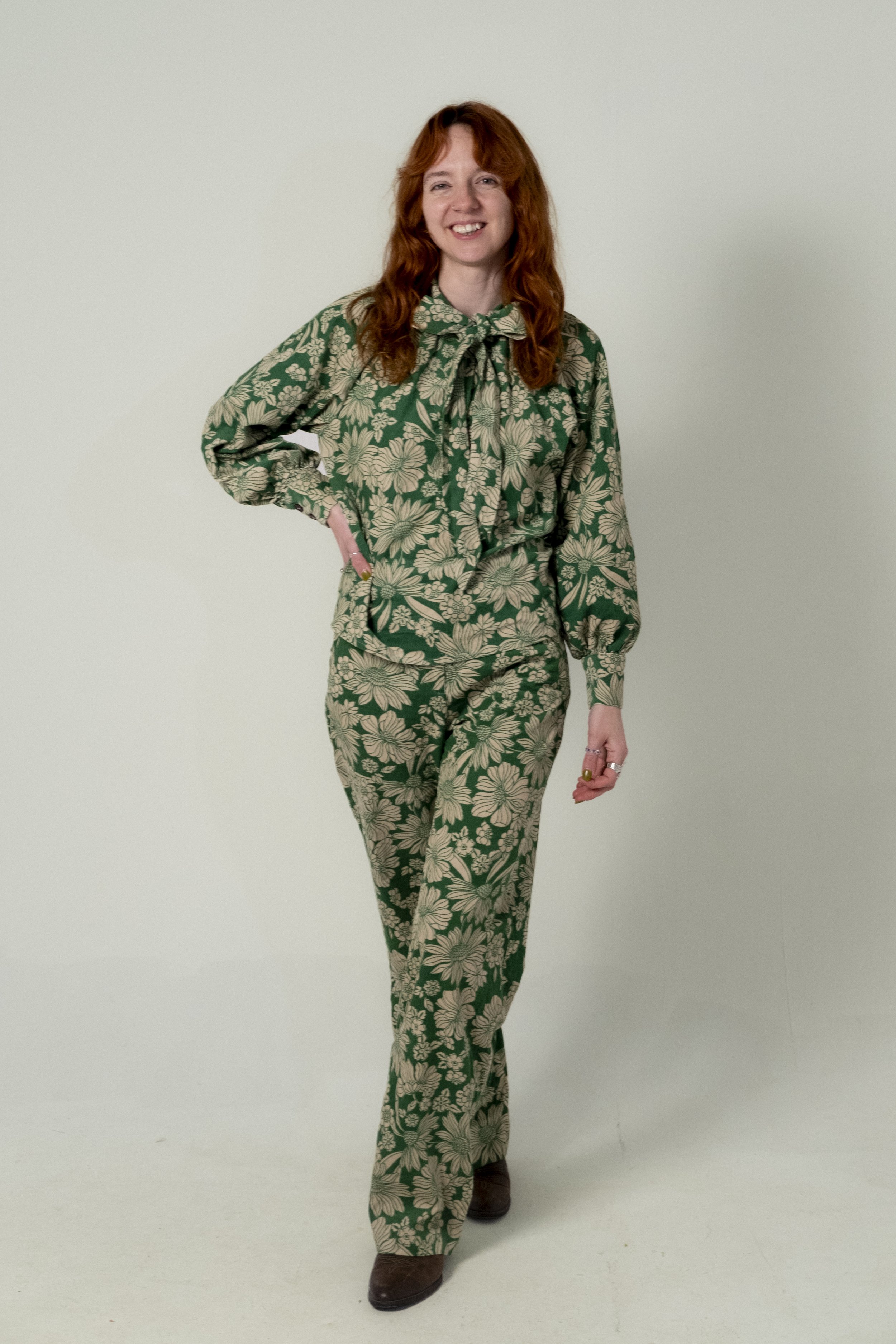 A woman with long red hair wearing a matching floral patterned green and beige outfit, standing against a plain light-colored background, smiling and posing with one hand on her hip.