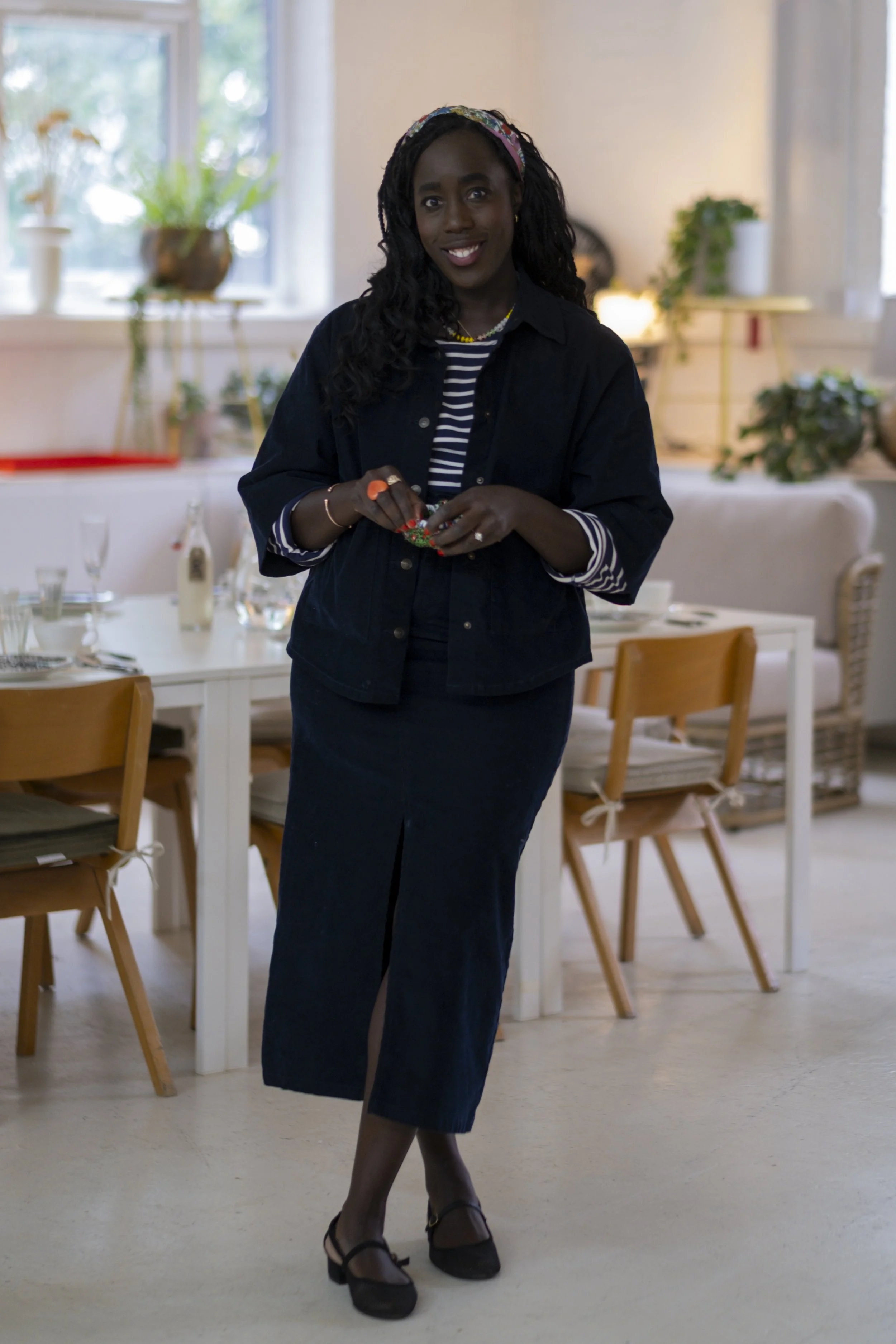 A woman with dark curly hair wearing a headband, a striped shirt, a dark jacket, and a long skirt standing in a well-lit dining room with wooden chairs and a table set for a meal. She is smiling and holding a small gift or ornament.