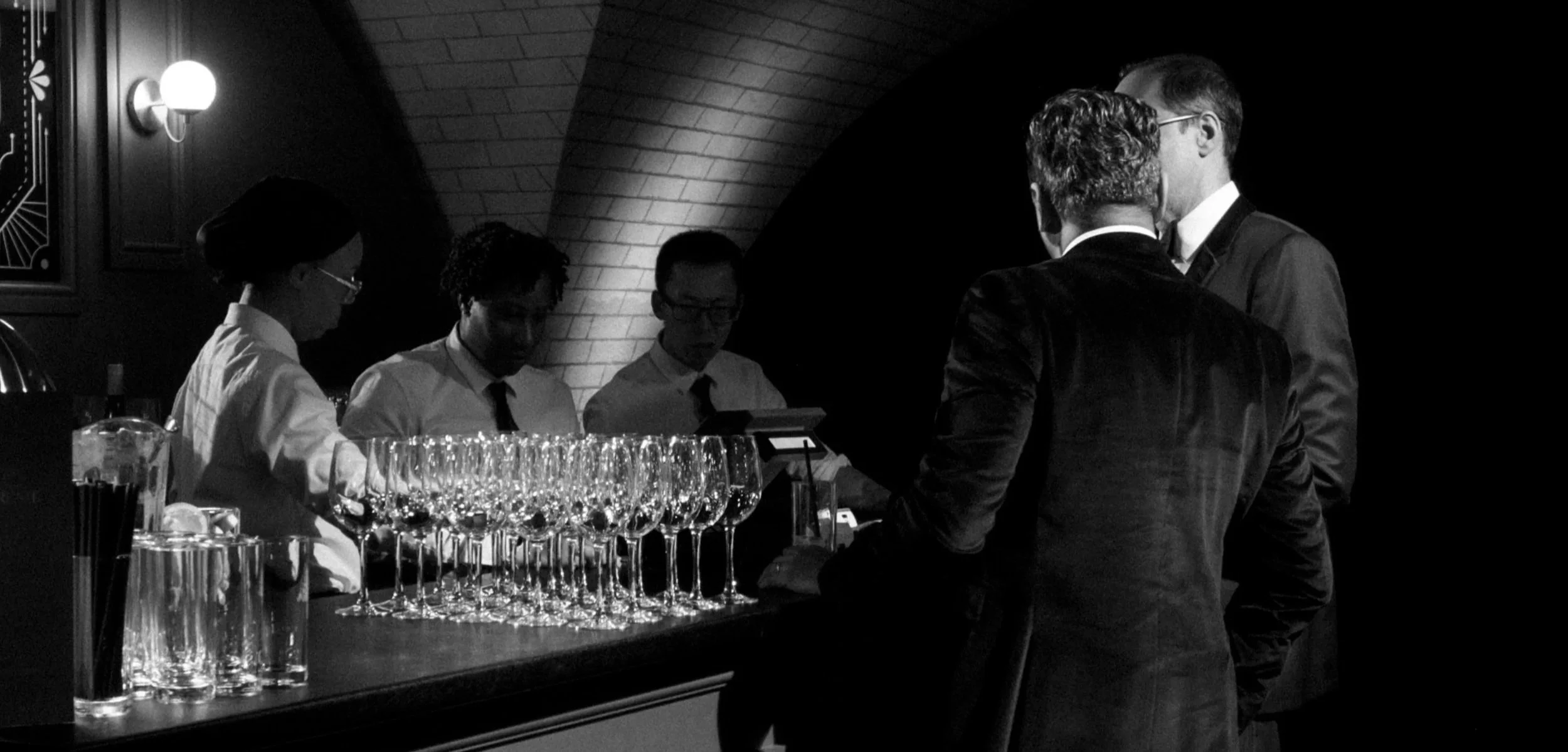 A black and white photo of a bar with four people, three behind the bar and two in front. The people behind the bar, presumably bartenders, are wearing white shirts and black ties, surrounded by wine glasses and other barware. The people in front are engaged in conversation, dressed in formal suits, with one man facing away and the other partially visible.