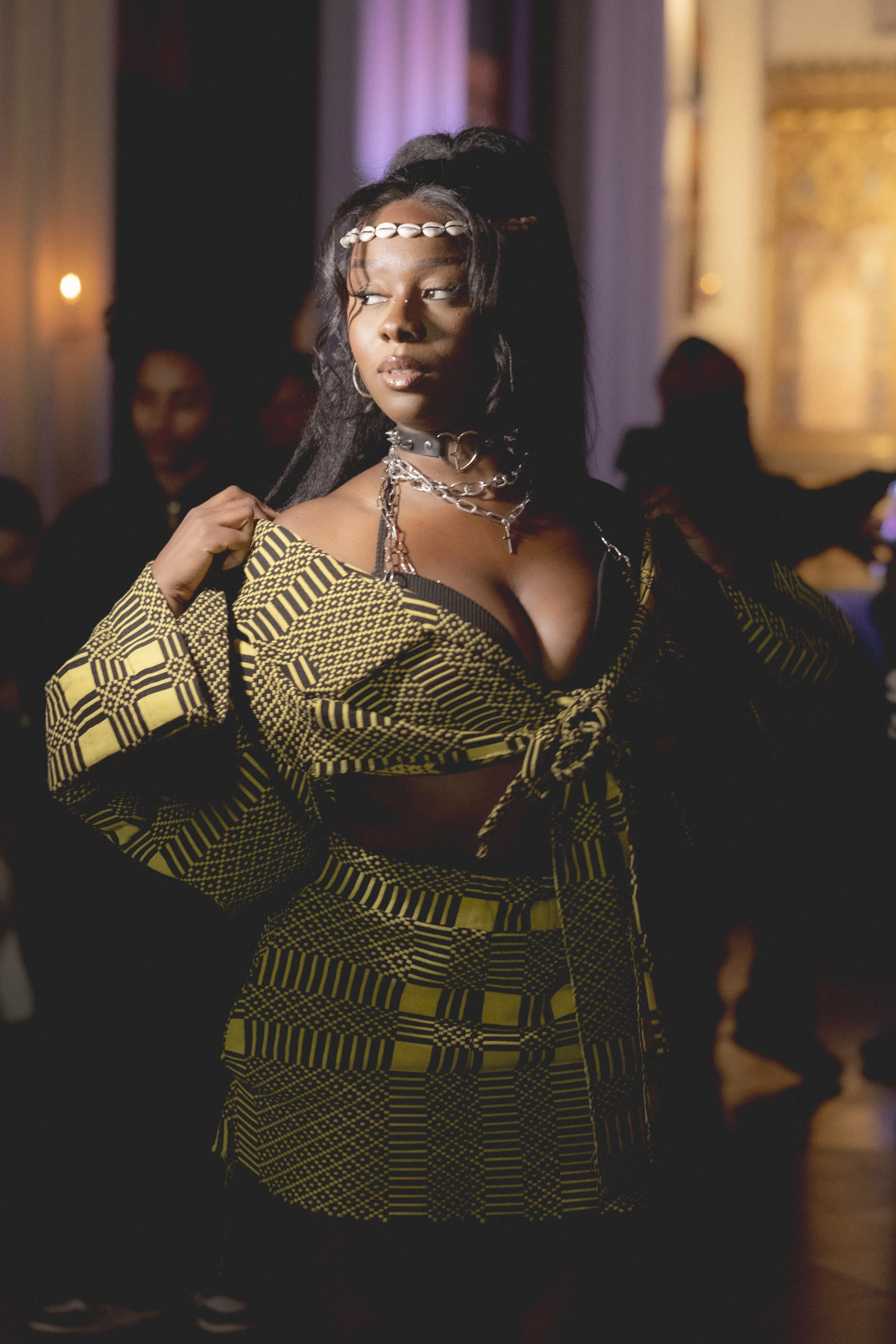 A woman with dark hair styled in waves, wearing a shell headband, silver jewelry, and a yellow and black patterned outfit, standing in a dimly lit room with other people in the background.
