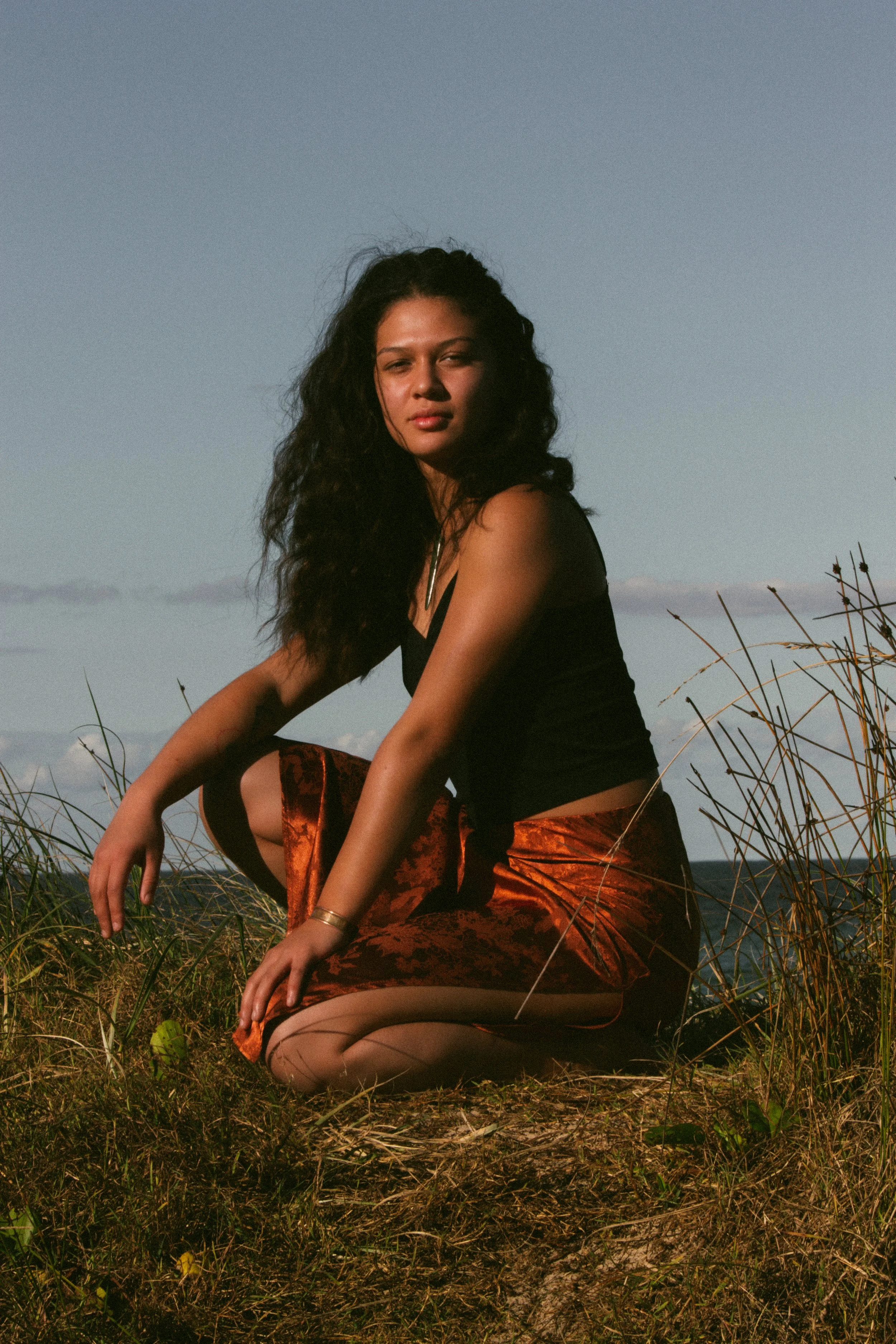 A woman with long, curly hair squatting on grassy ground outdoors, wearing a black top and patterned skirt, with a blue sky and distant landscape in the background.
