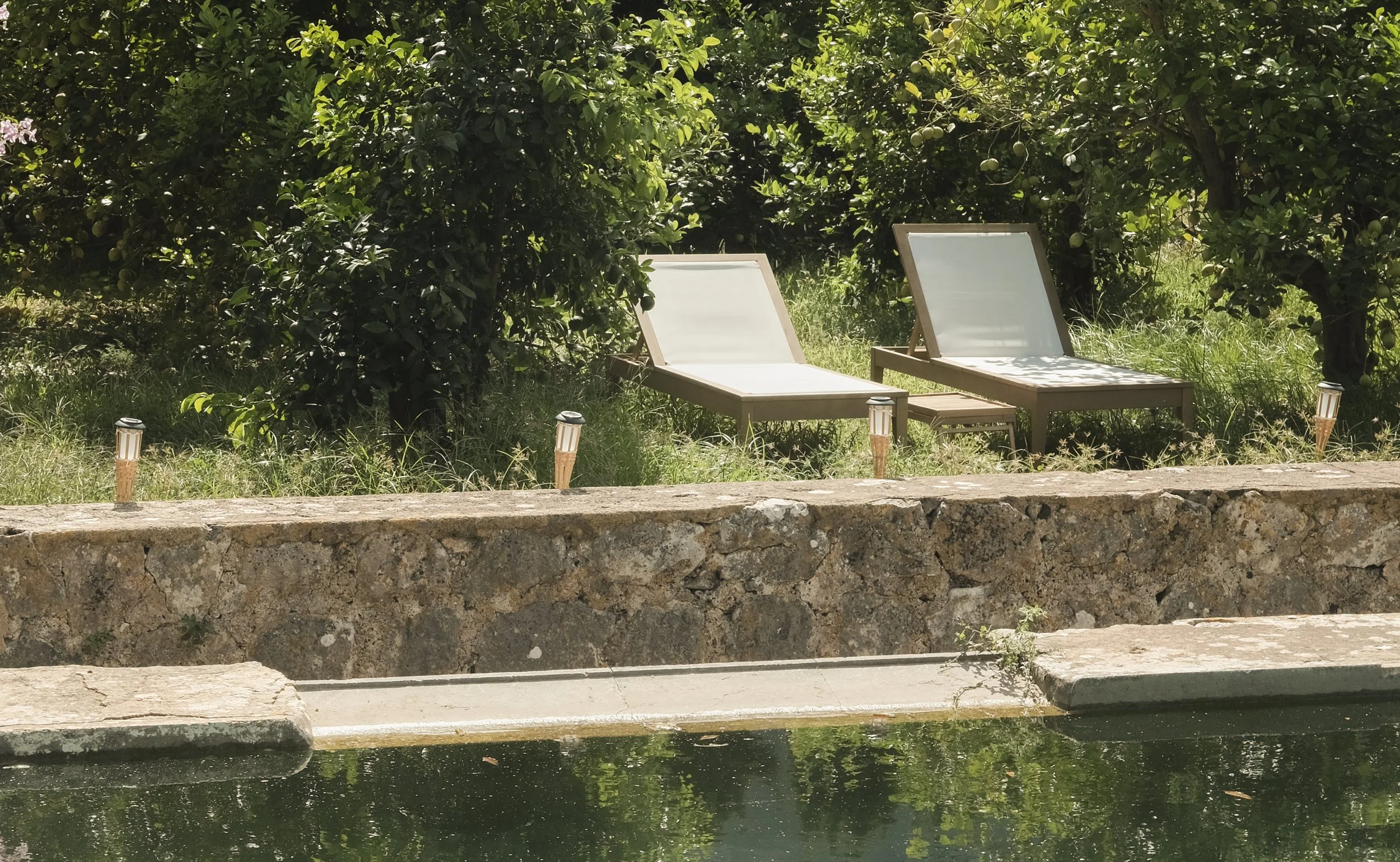 Two white lounge chairs are placed outside amidst green foliage, with a stone wall and a body of water in the foreground.