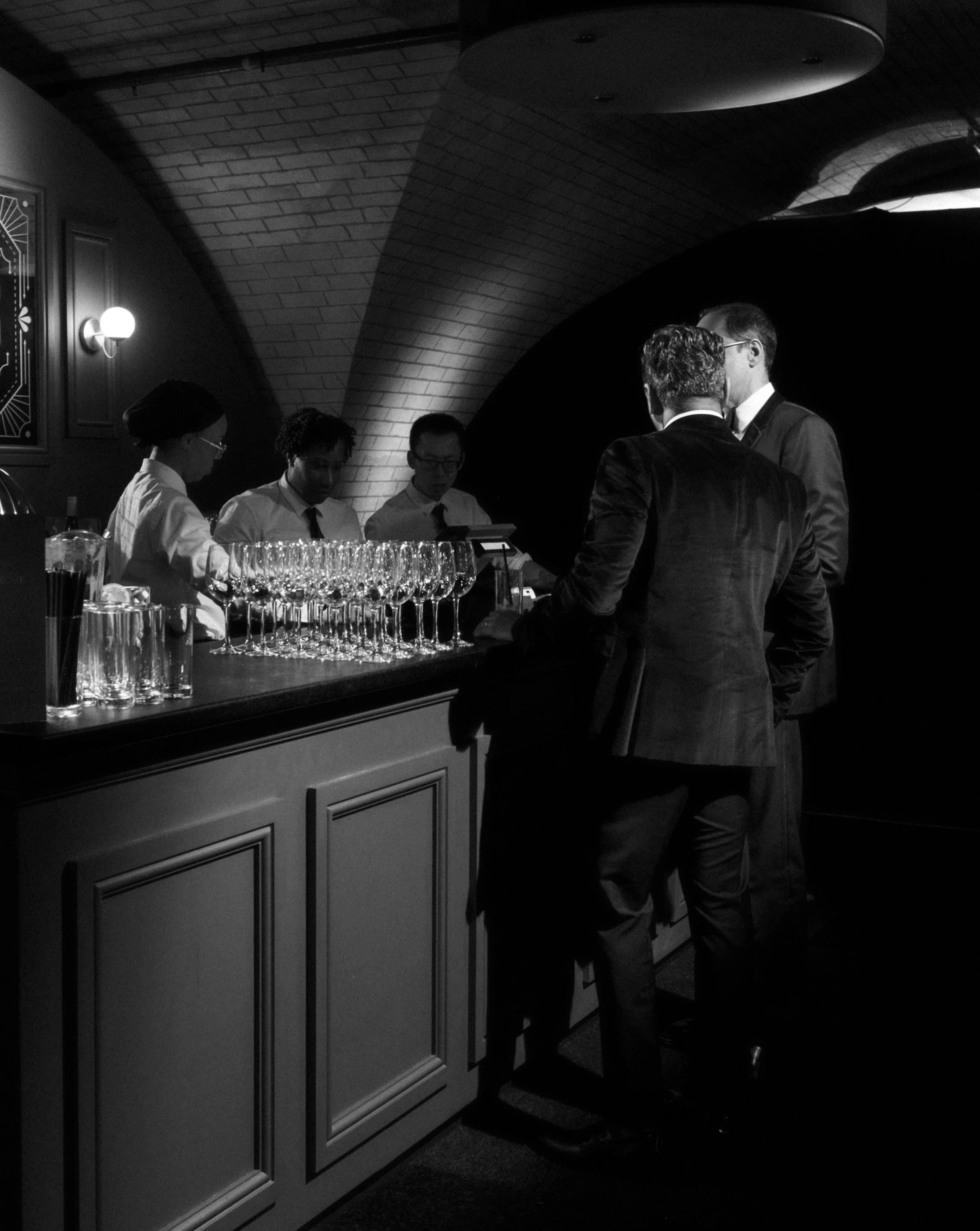 People gathered around a bar counter in a dimly lit, stylish venue with a brick arch ceiling, engaging in conversation.