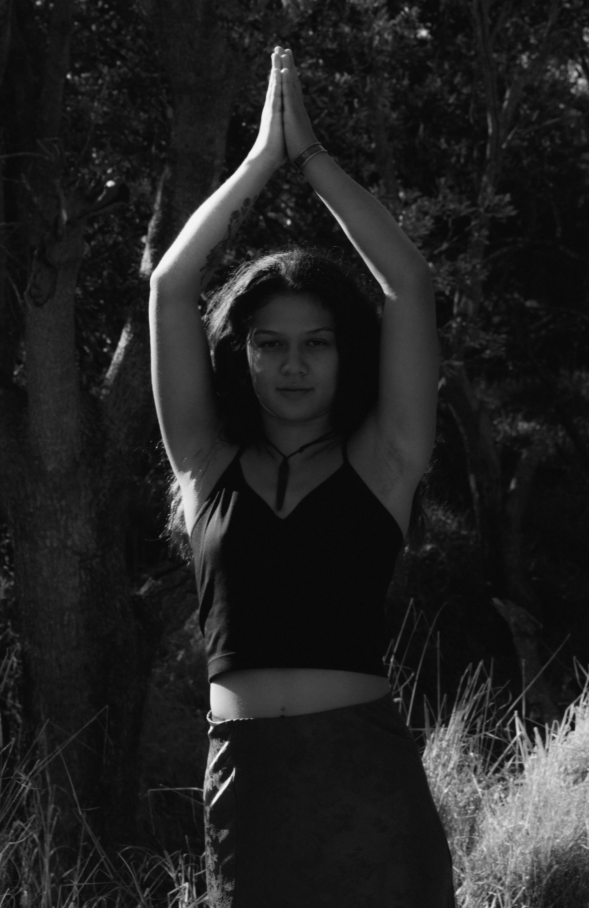 A woman standing outdoors with her arms raised above her head in a prayer-like pose. She is facing the camera, with a slight smile, and dark, wavy hair. The background features trees and grassy terrain, and the photo is in black and white.