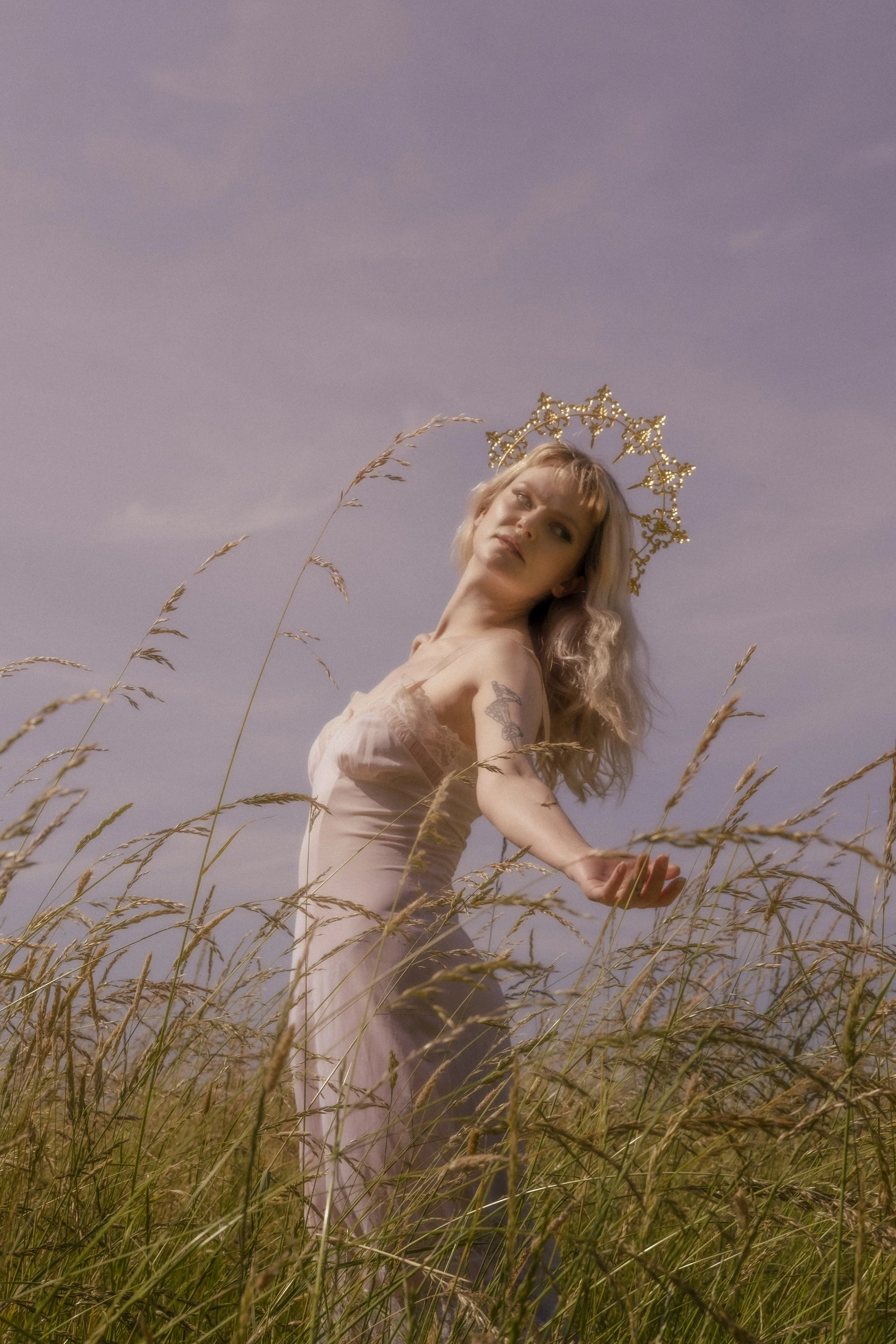 A woman with blonde wavy hair standing in a field of tall, dry grass, wearing a light-colored dress and a decorative gold crown, with a peaceful expression and a slight tilt of her head against a sky with soft clouds.
