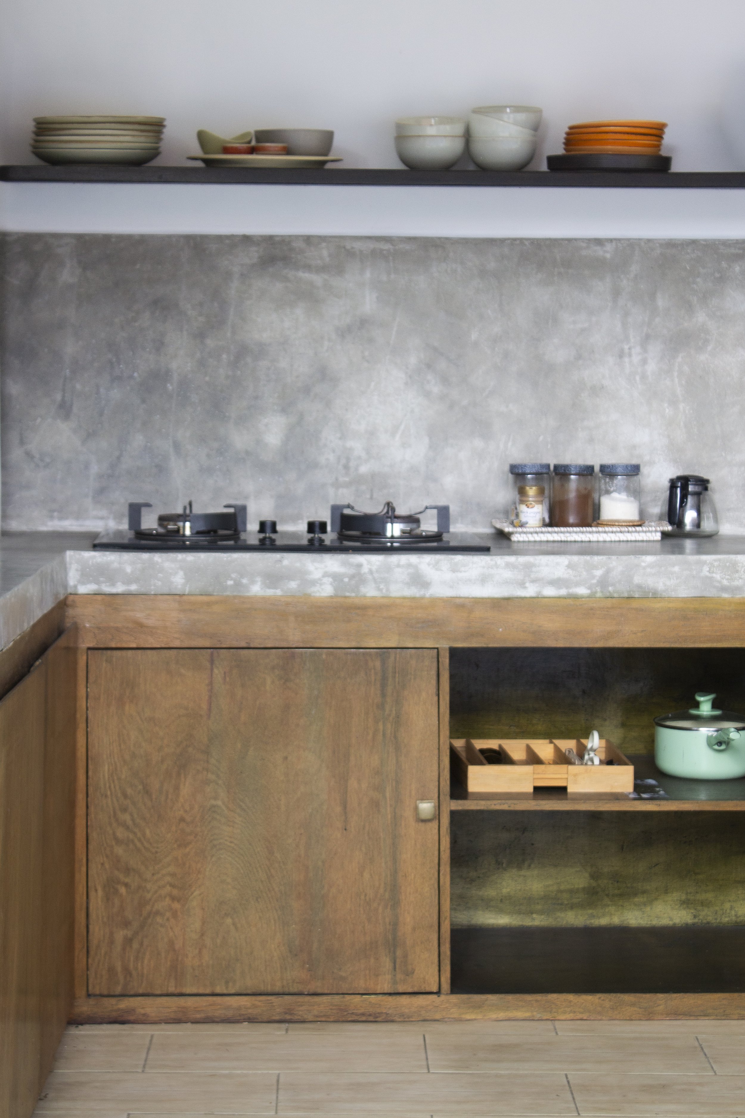 Kitchen counter with wooden cabinetry, a sink with a faucet, and a teal pot, with open shelves above holding bowls, plates, spices, and other kitchen items.