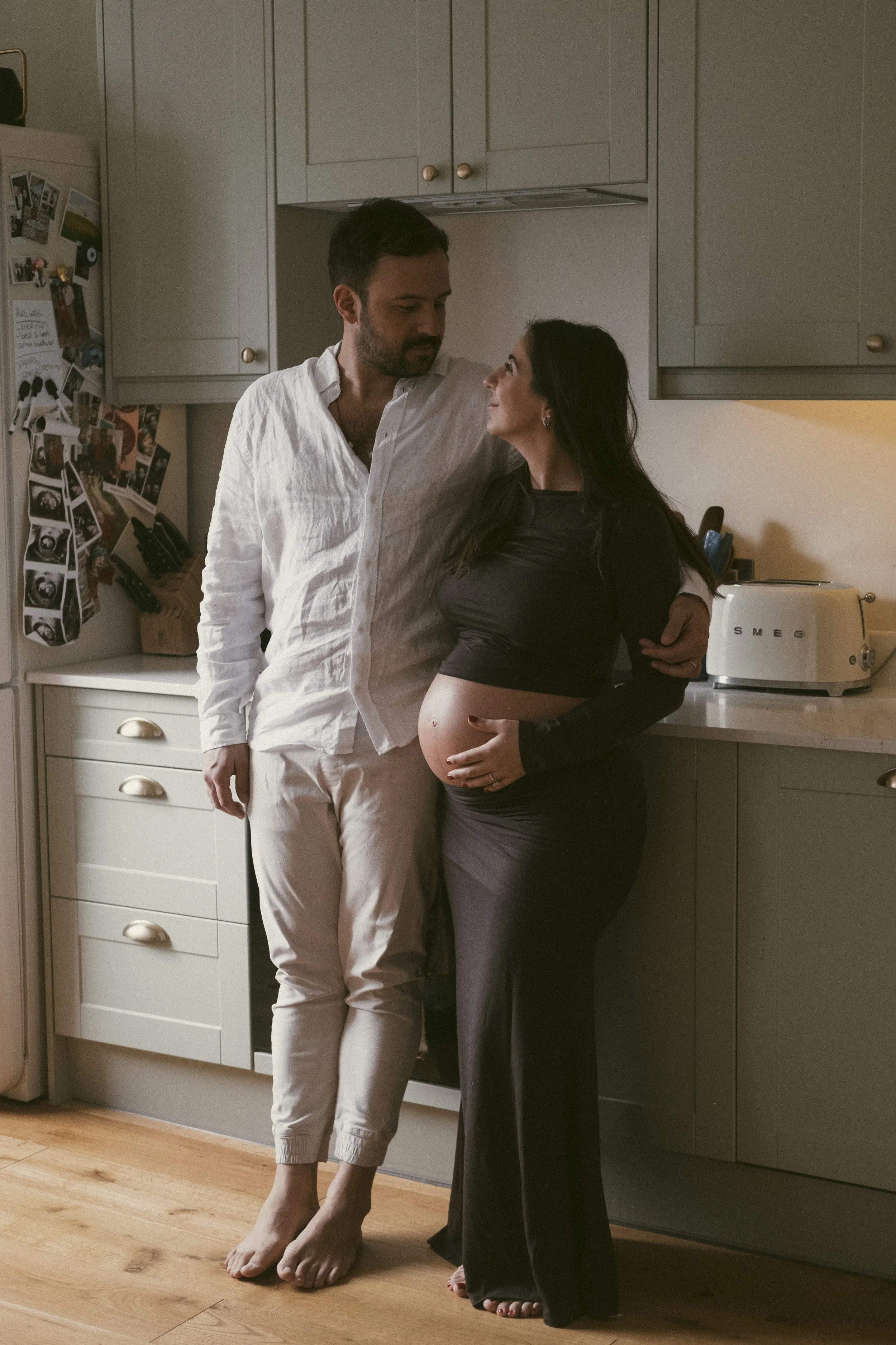 A pregnant woman stands in a kitchen, holding her belly and looking at her partner, while a man stands beside her, looking at her affectionately.