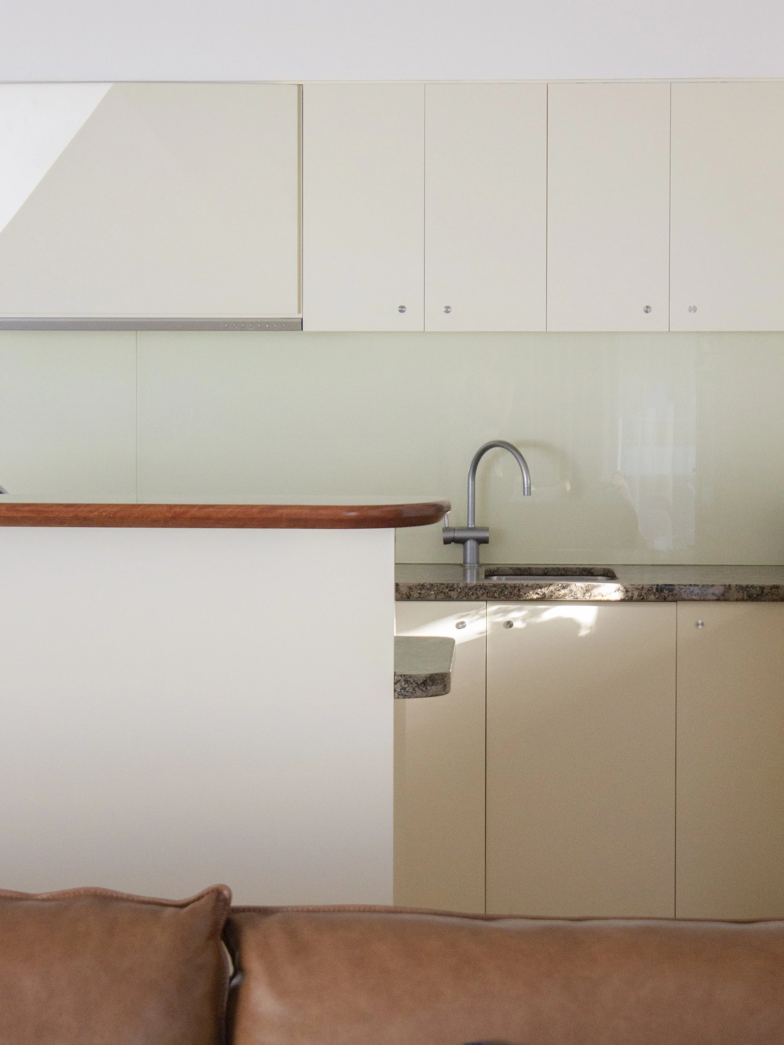 An interior view of a kitchen with white cabinets, a granite countertop, a sink with a silver faucet, and a brown sofa or chair in the foreground.