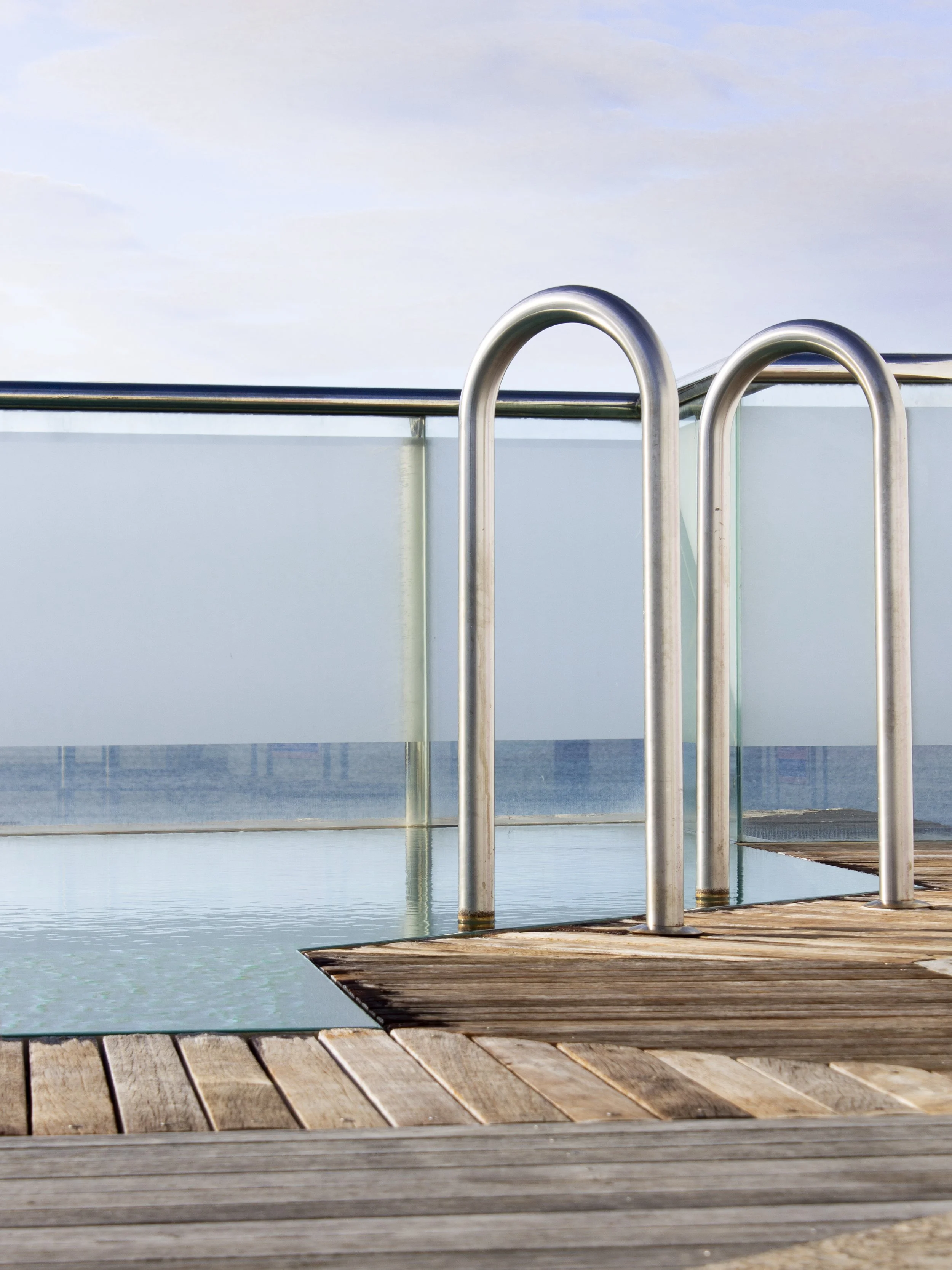 Swimming pool with metal ladder, wooden deck, and glass barrier, with cloudy sky in background.