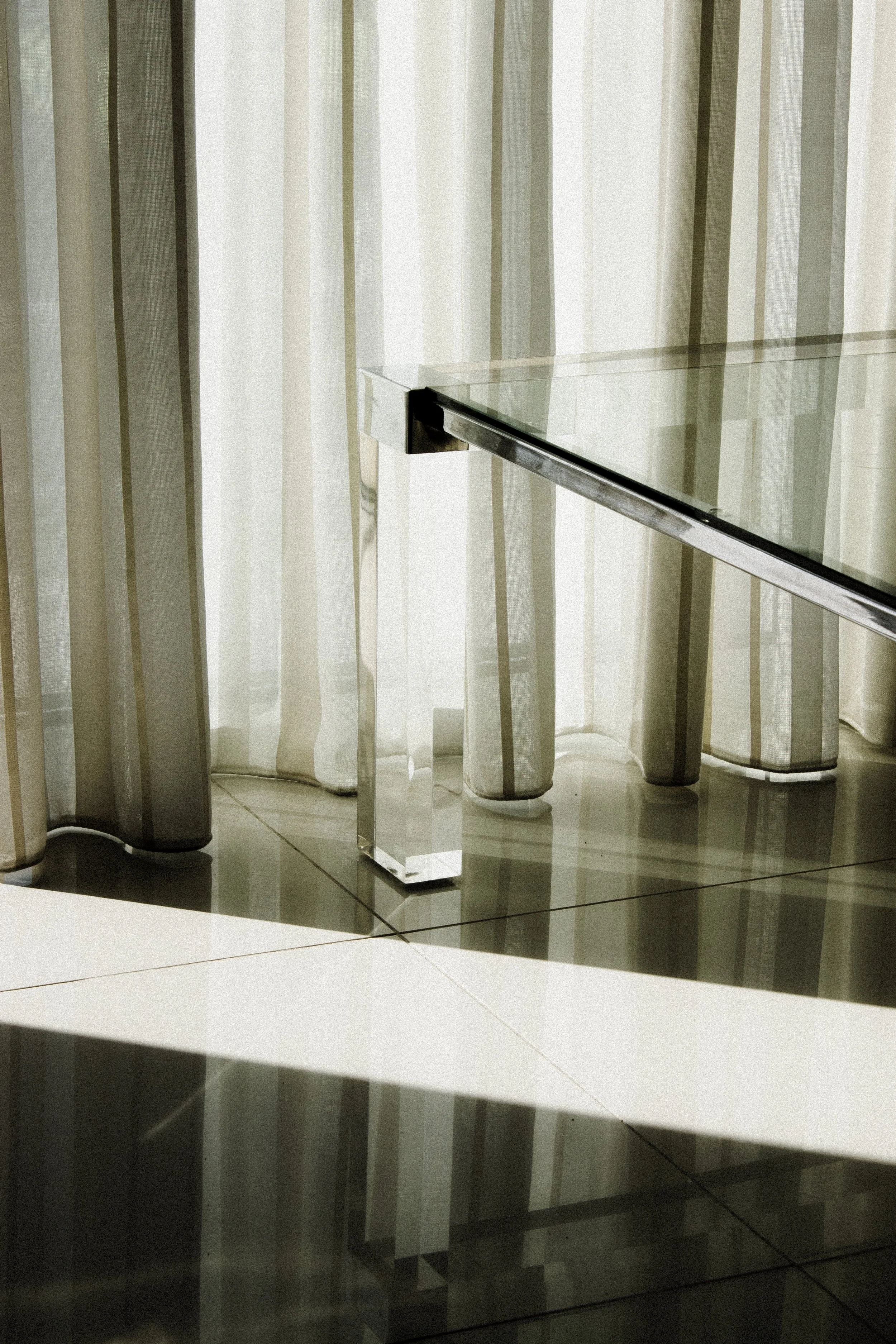 A modern glass and metal desk with a reflective black base, situated in front of sheer white and gray striped curtains, with a tiled floor beneath.