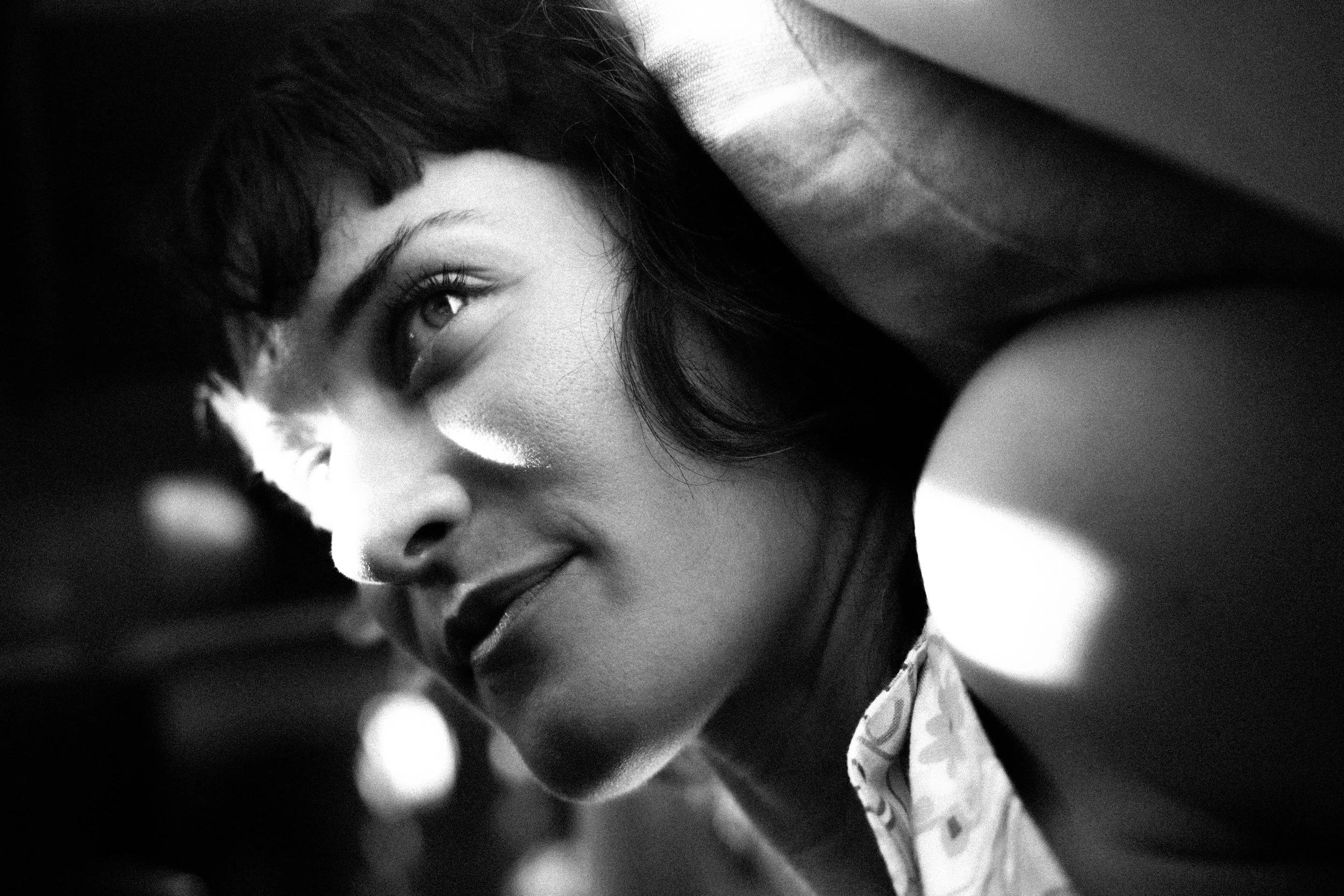 A black and white close-up photograph of a young woman with dark hair, smiling with her eye closed, wearing a patterned shirt.