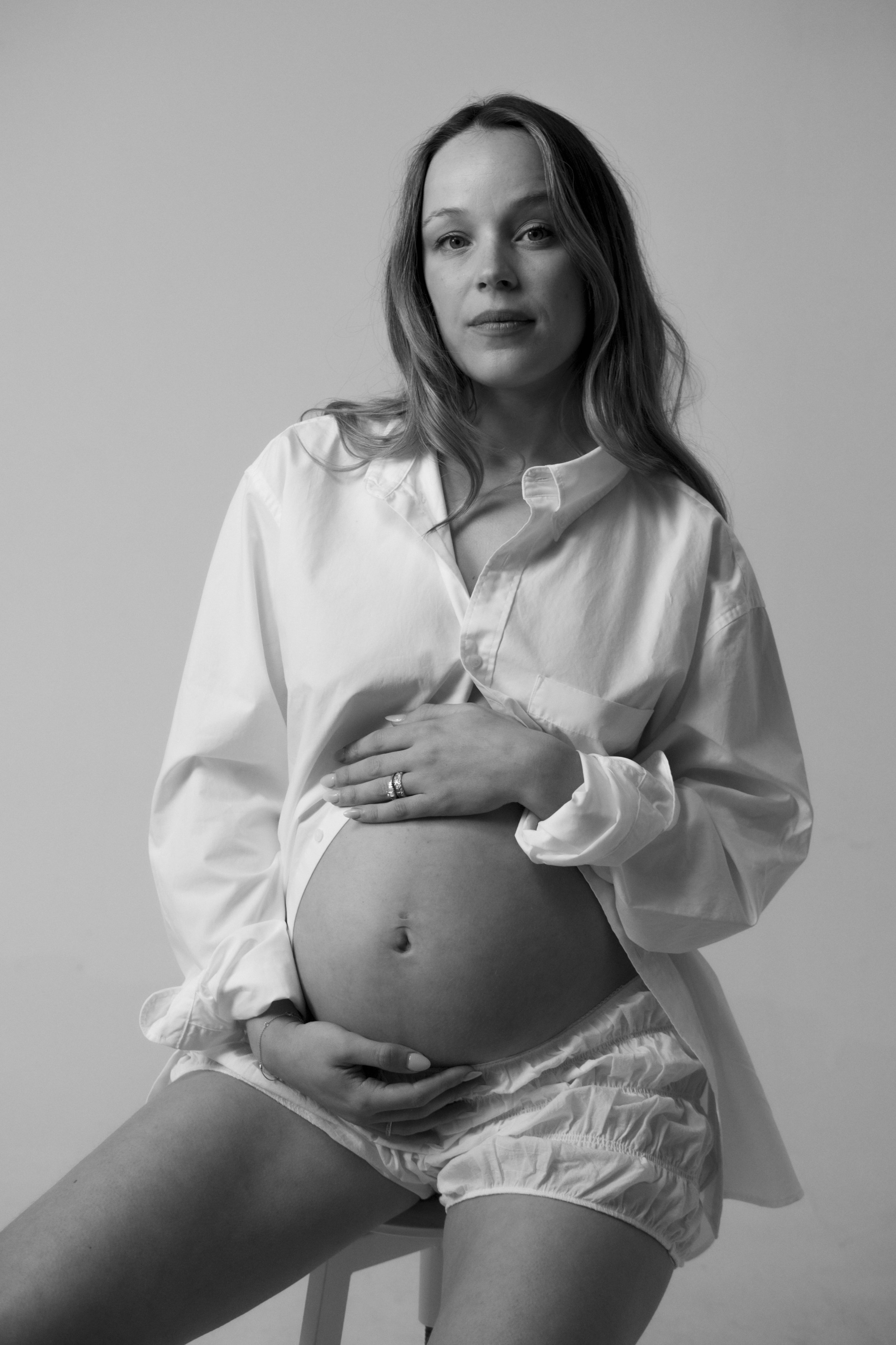 A black-and-white portrait of a pregnant woman wearing an open white shirt and shorts, sitting on a chair, with her hand resting on her belly.